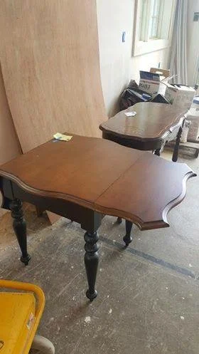 Two wooden tables, one with curved edges in the foreground and another smaller one in the background, in an unfinished room with a window and various boxes and items around.