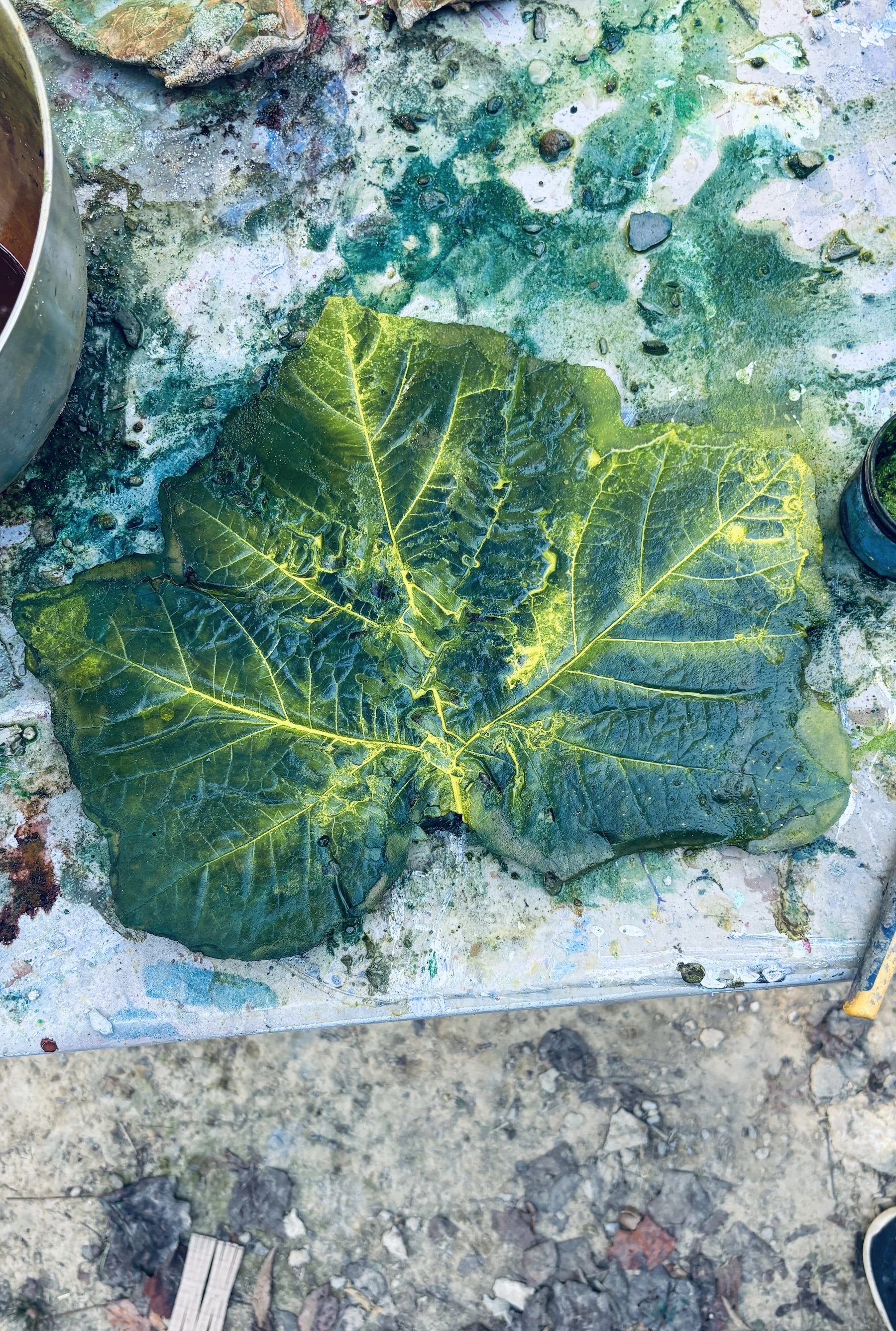 A large, green, richly veined ceramic leaf on a messy, colorful workshop table with paint and water stains.