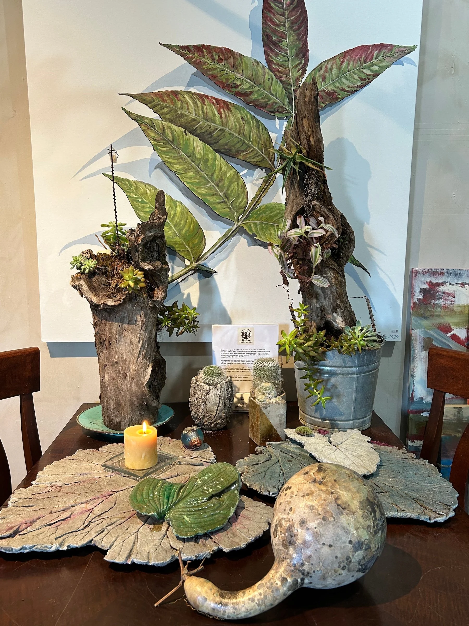 Decorative display featuring various plants, cacti, a candle, and natural objects on a wooden table, with a botanical artwork in the background.