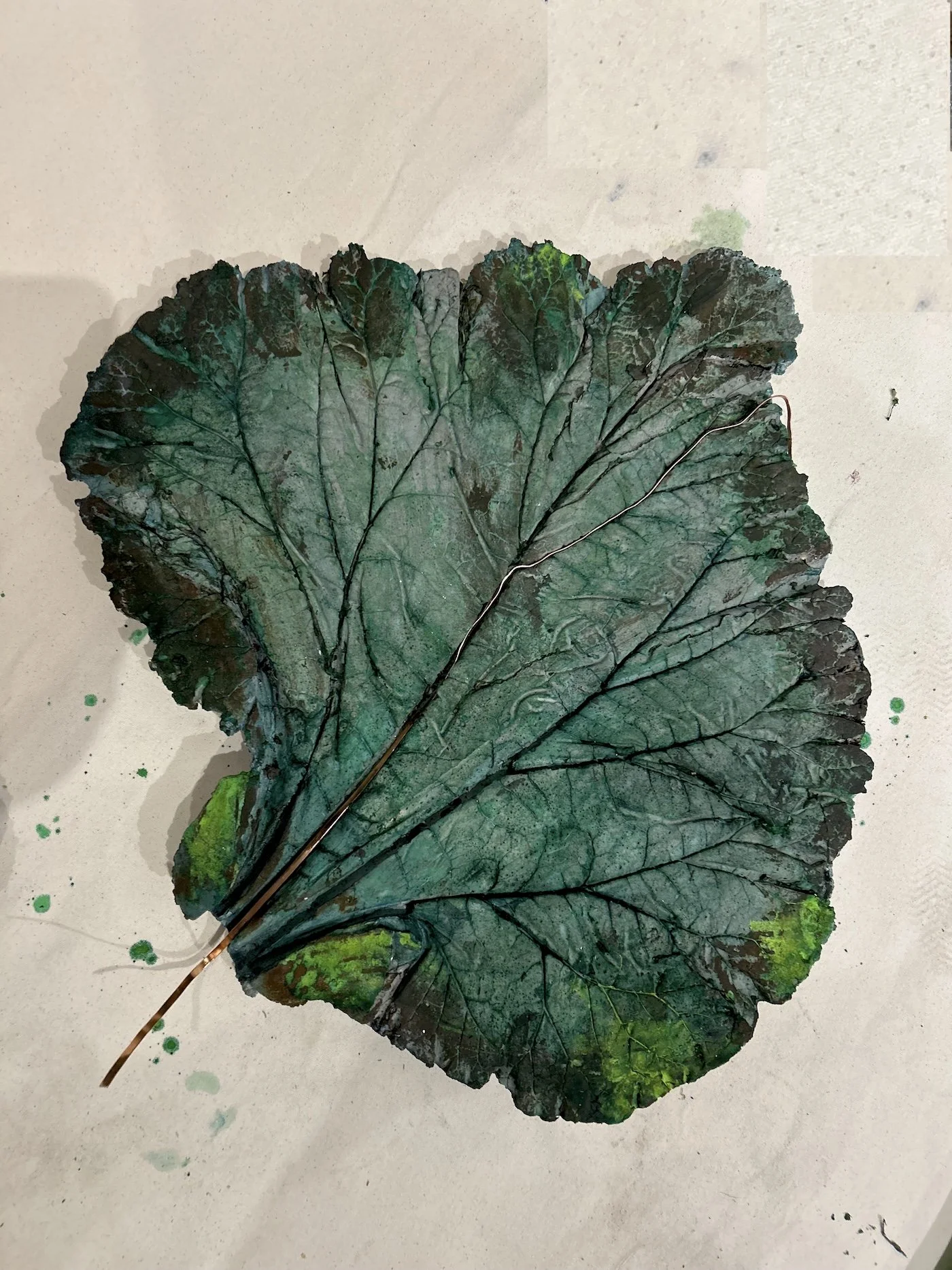 A dried green ceramic leaf with dark veins and a long, thin stem, placed on a light-colored surface with some green and black paint splatters.