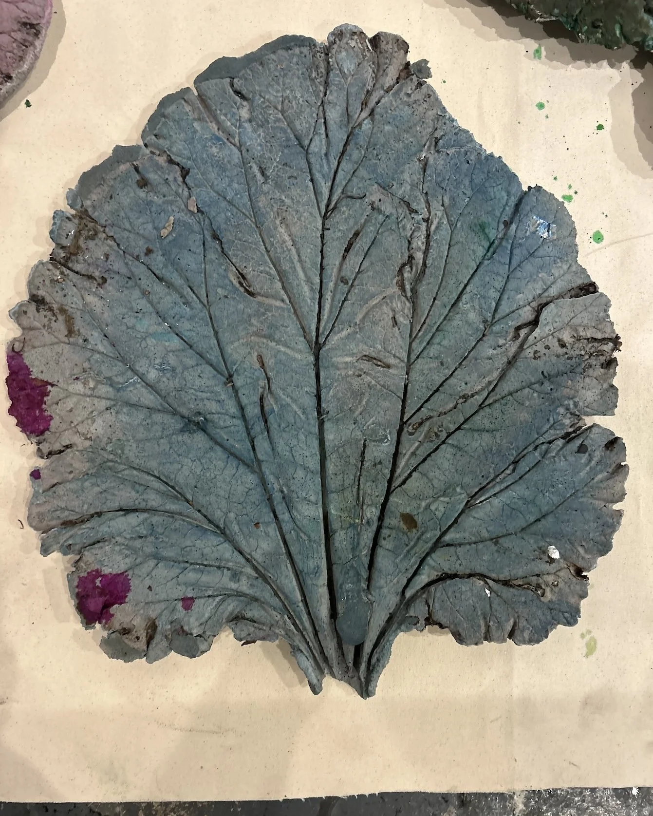 A large, dried, blue-gray ceramic leaf with prominent veins and slightly frayed edges, placed on a light-colored surface with small green and pink paint splatters around it.