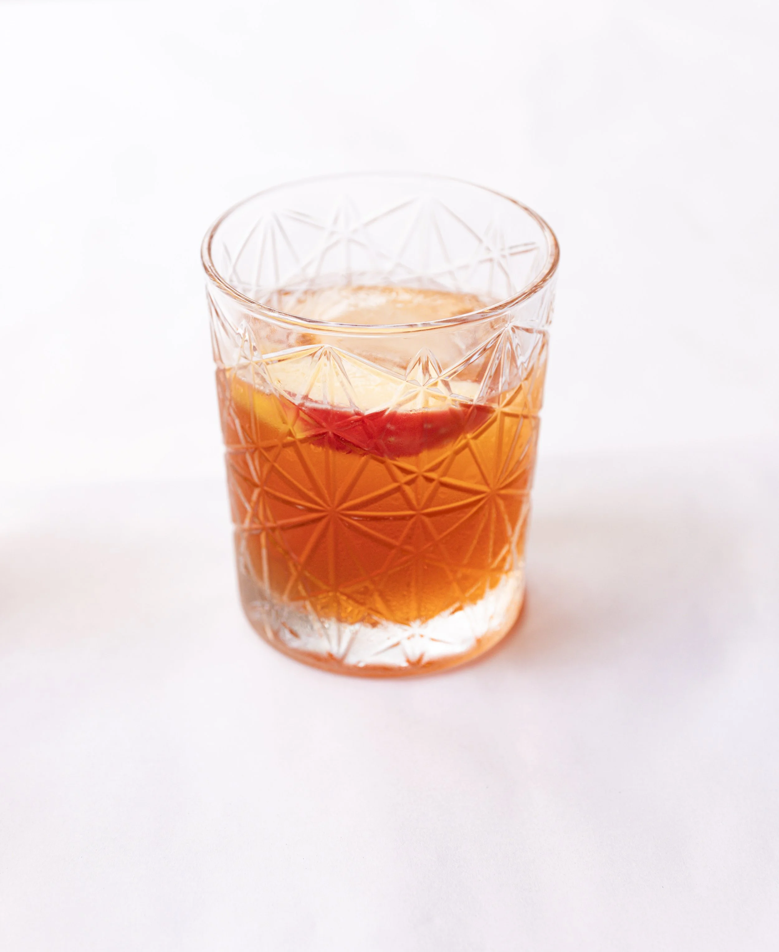 A glass of orange-colored cocktail with a lemon wedge and a cherry garnish on top.