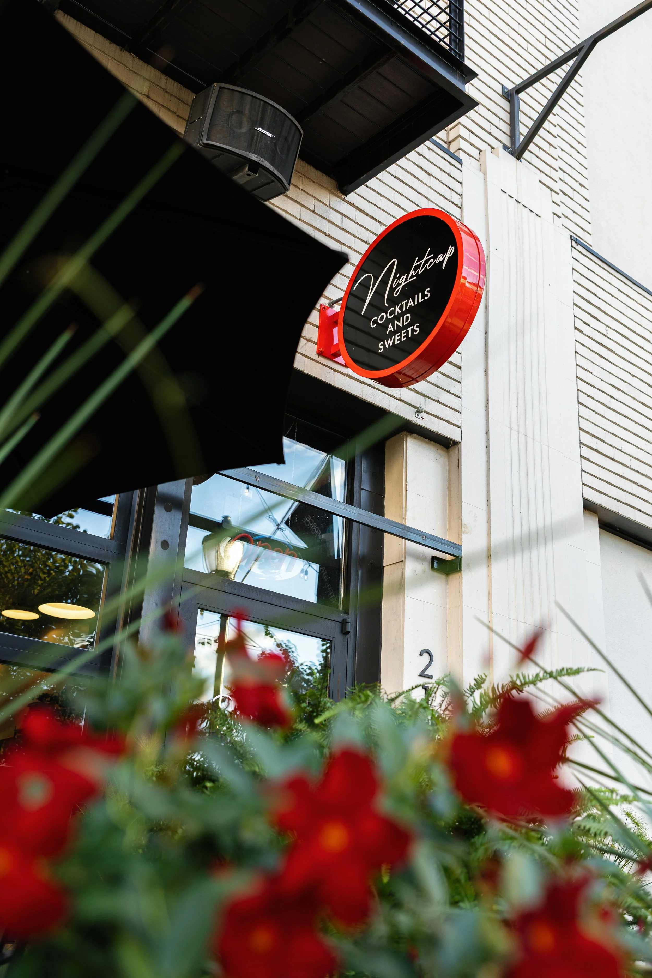Exterior view of a café or bar named Nightcap with a red circular sign that reads 'Nightcap Cocktails and Sweets' mounted on a building with brick and white siding. In the foreground, there are red flowers and green foliage, slightly out of focus.
