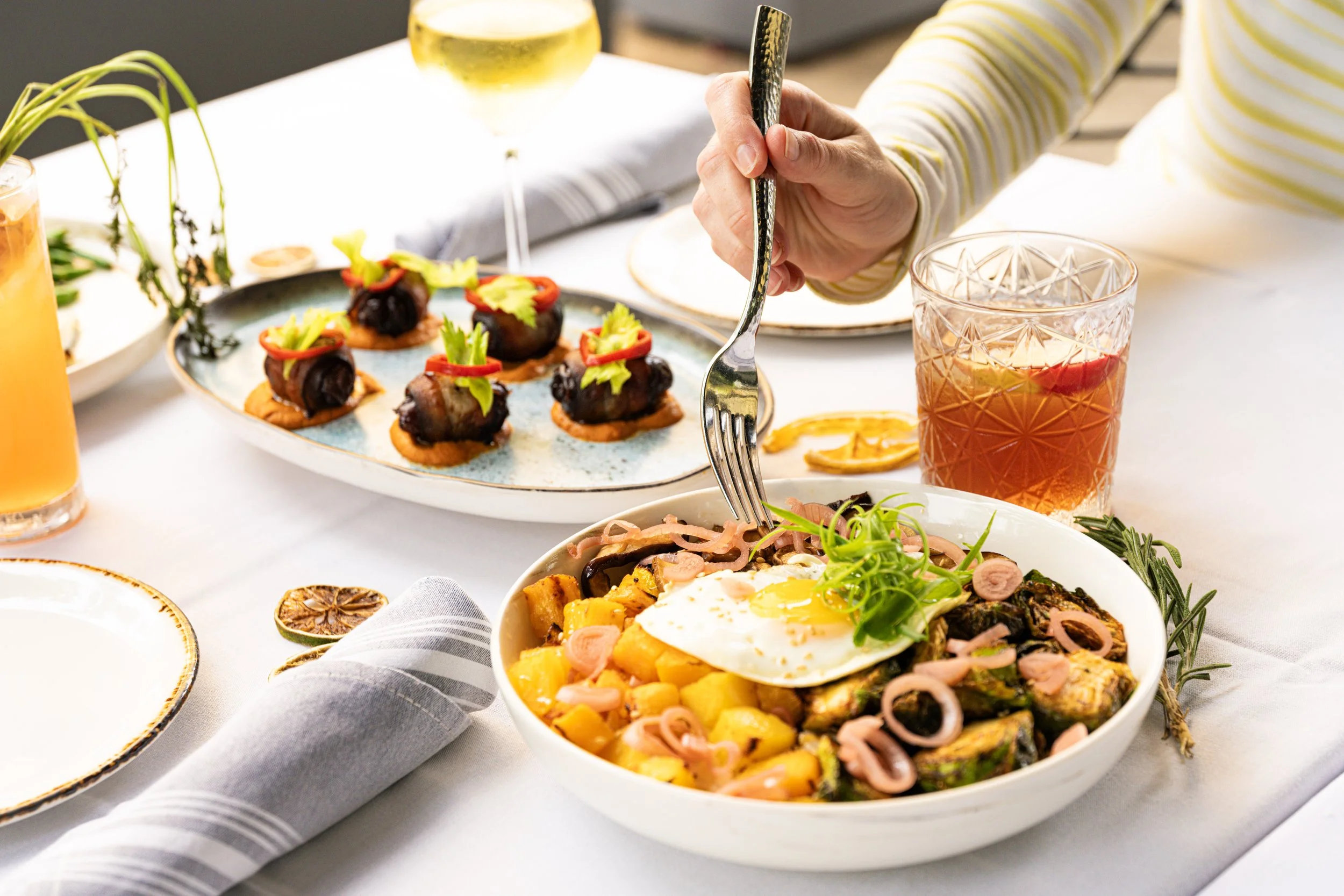 A person is about to eat a bowl of roasted vegetables and a fried egg, with a plate of appetizers and drinks on a white table.