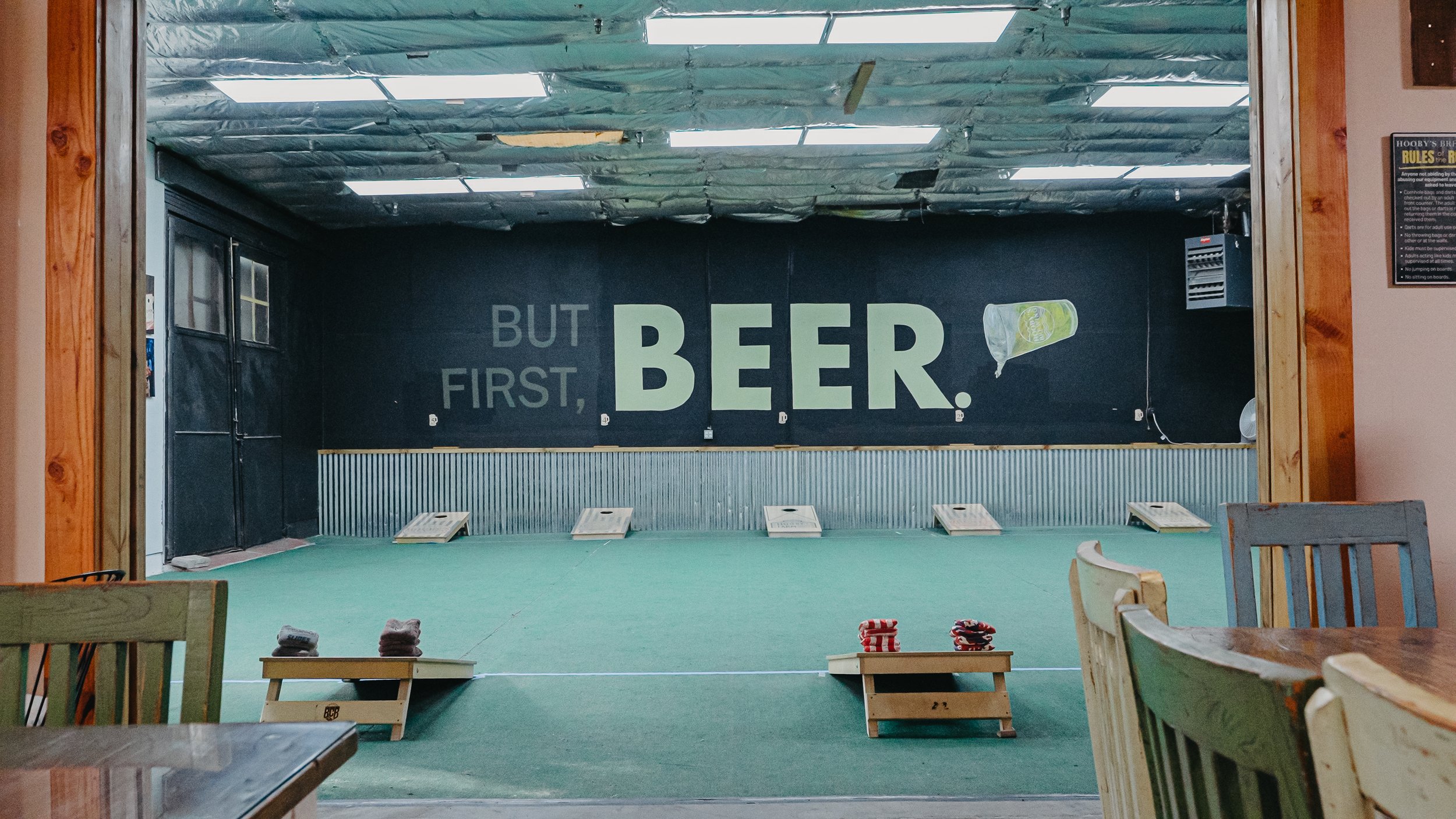 a room with cornhole boards with the wall reading but first beer