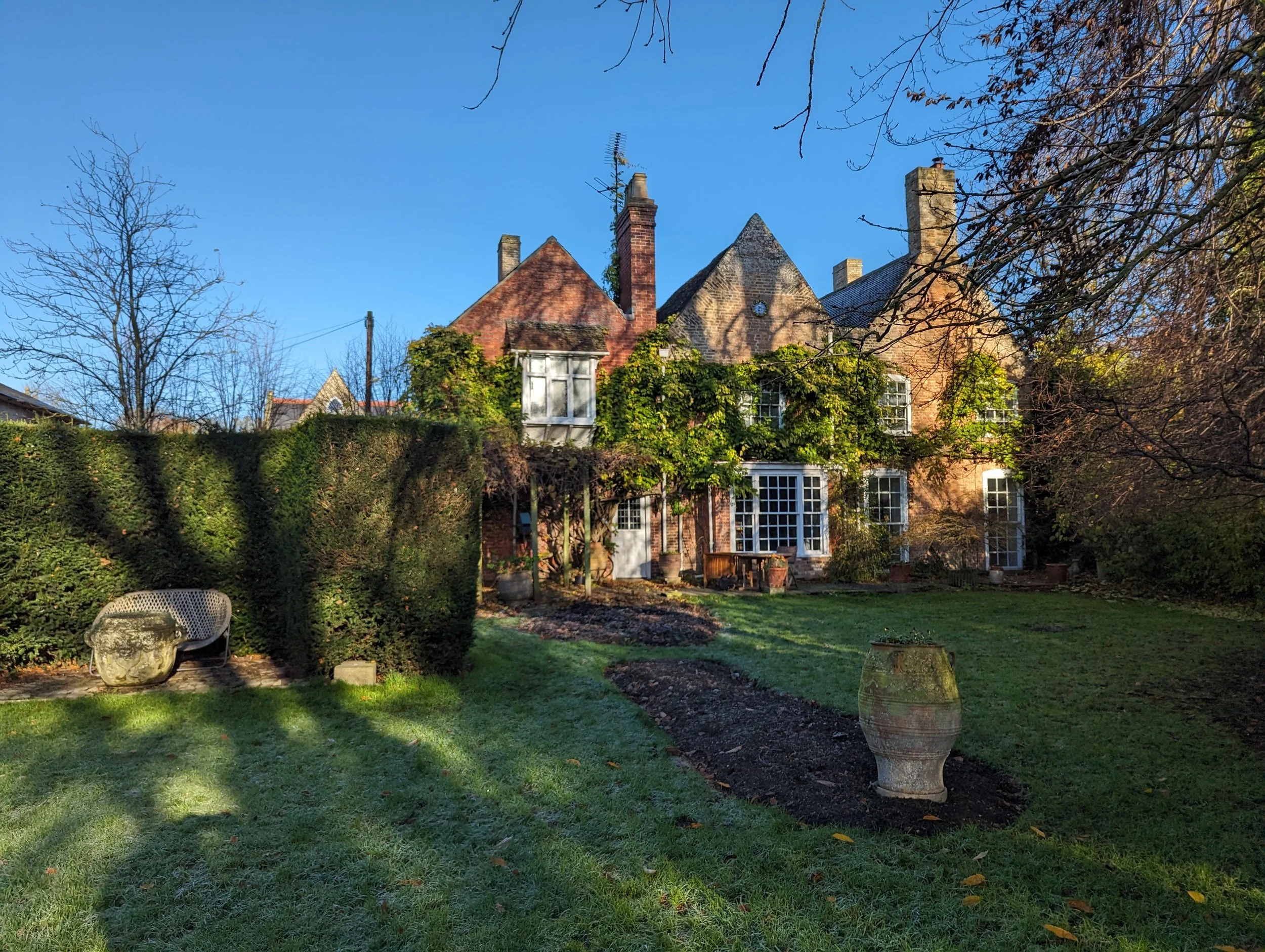 View of a cozy brick house with multiple chimneys and a sloped roof, surrounded by trees and lush greenery, with a grassy garden in the foreground and clear blue sky above.