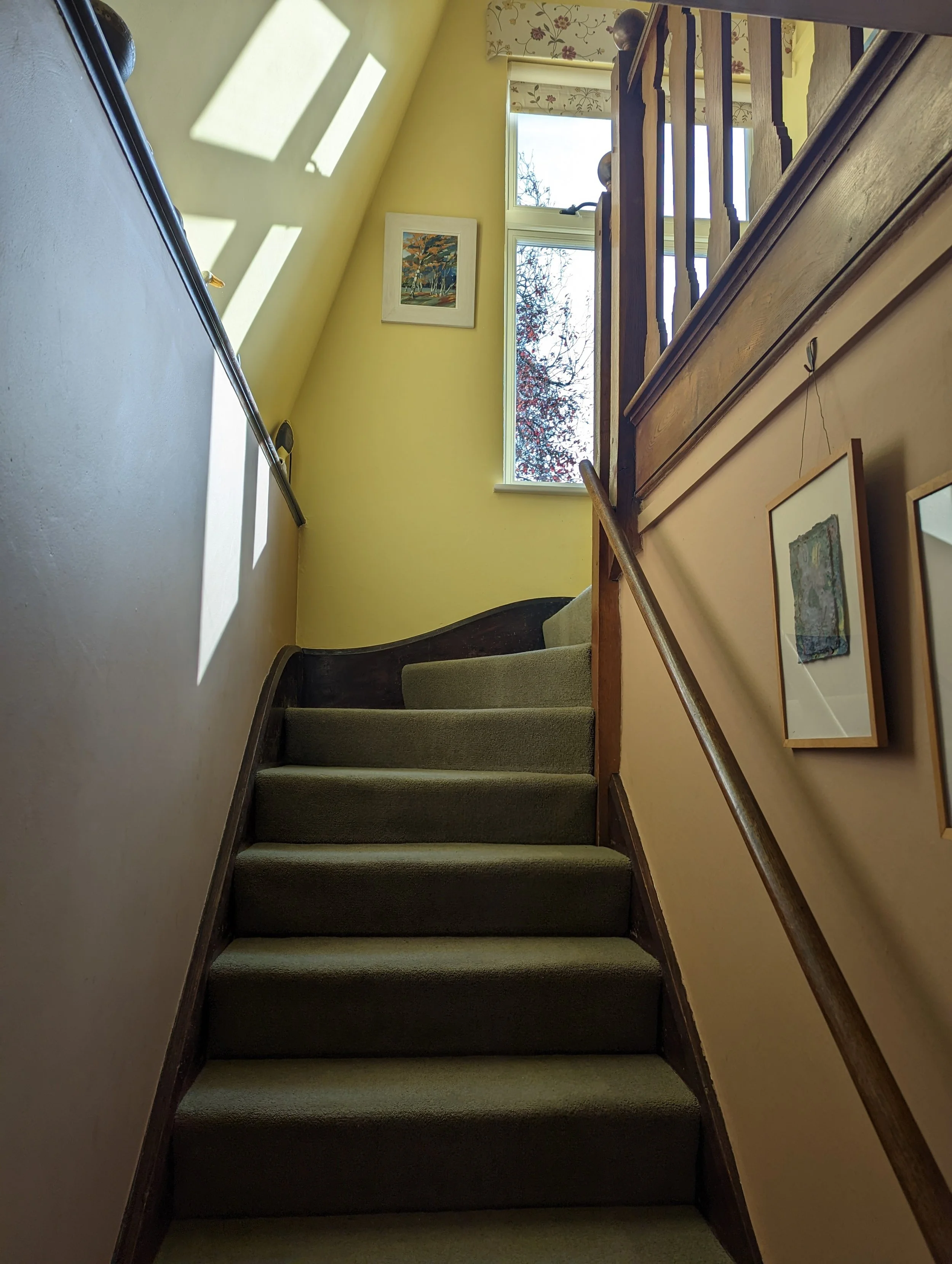 A staircase with green carpeted steps, wooden handrails, yellow walls, artwork on the walls, and windows letting in sunlight, with a view of trees outside.