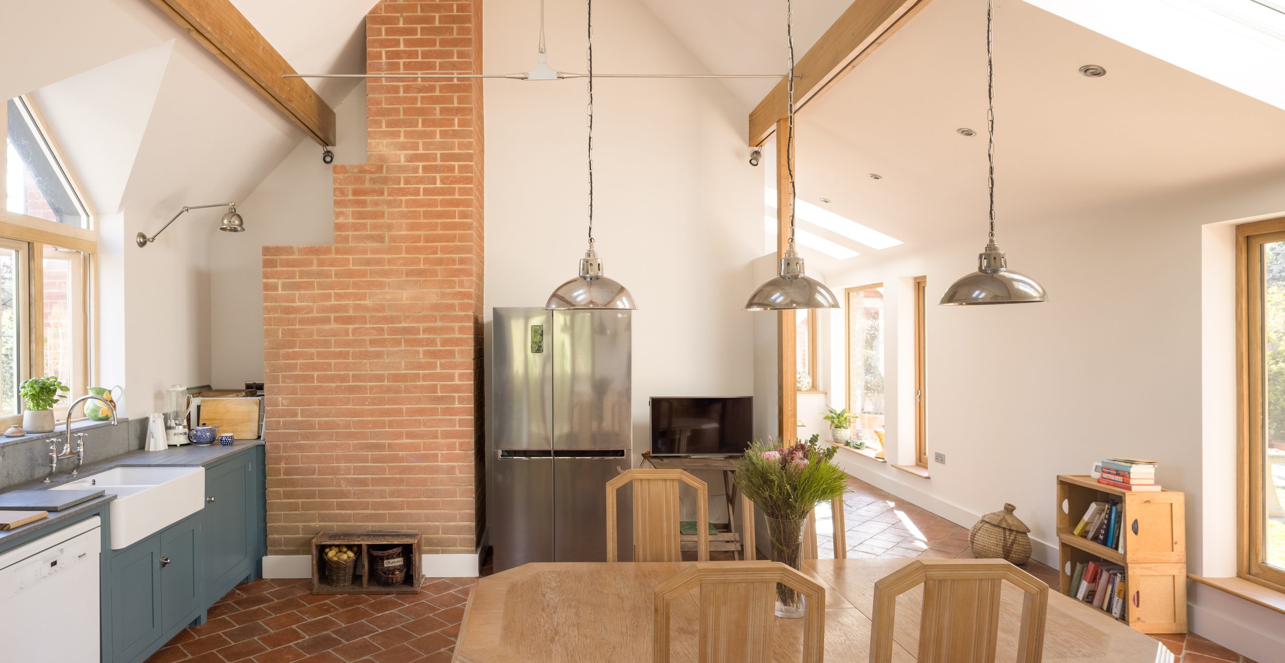 A bright, modern kitchen and dining area with large windows, a brick chimney, hanging pendant lights, a gray kitchen sink, and a wooden dining table with chairs.
