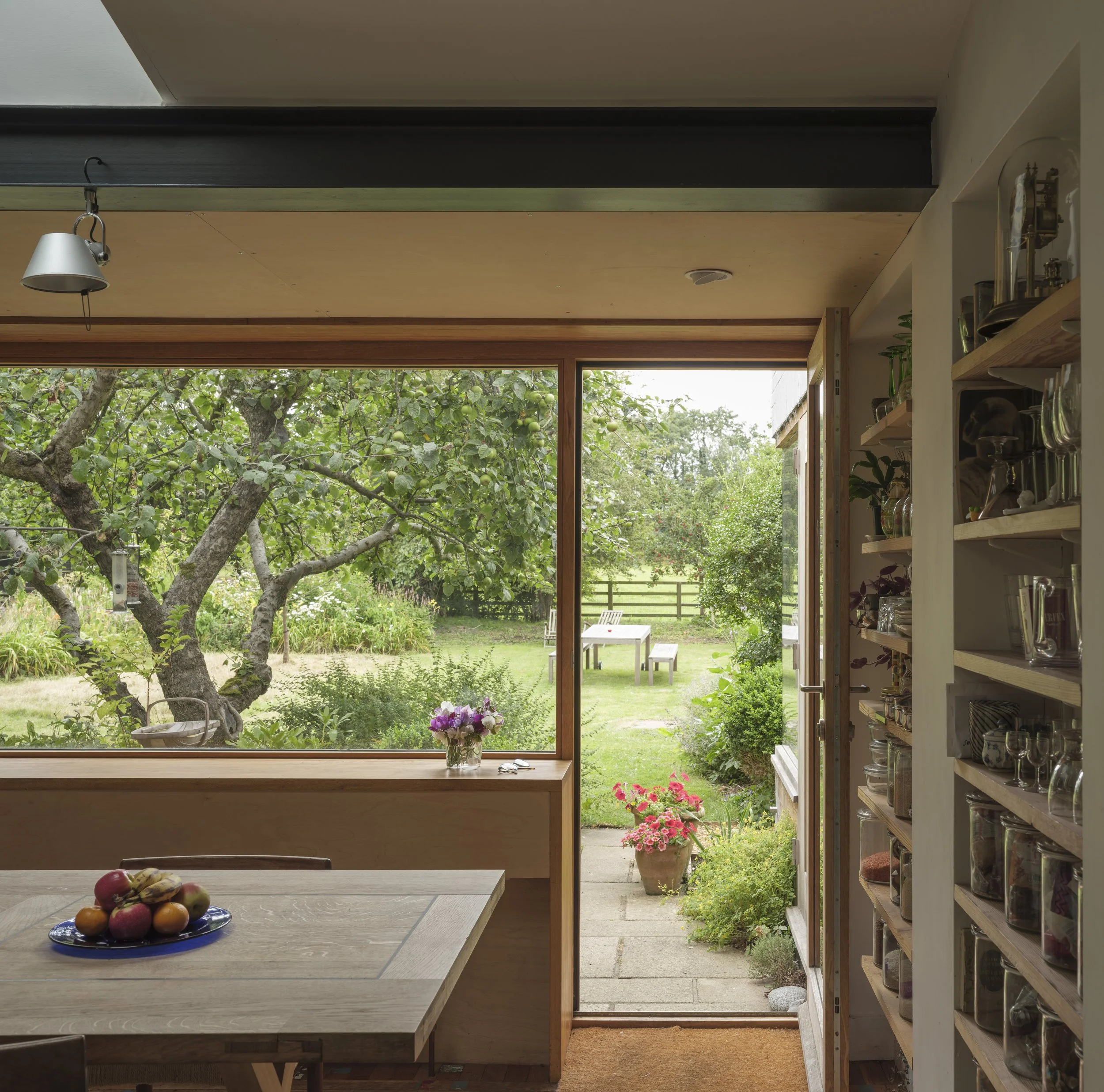 Interior view of a kitchen or dining area with large window showing a garden outside. A wooden table with a bowl of apples and bananas, and a vase of flowers. To the right, a shelf with glass jars and decorative items. Outside, there are trees, a law