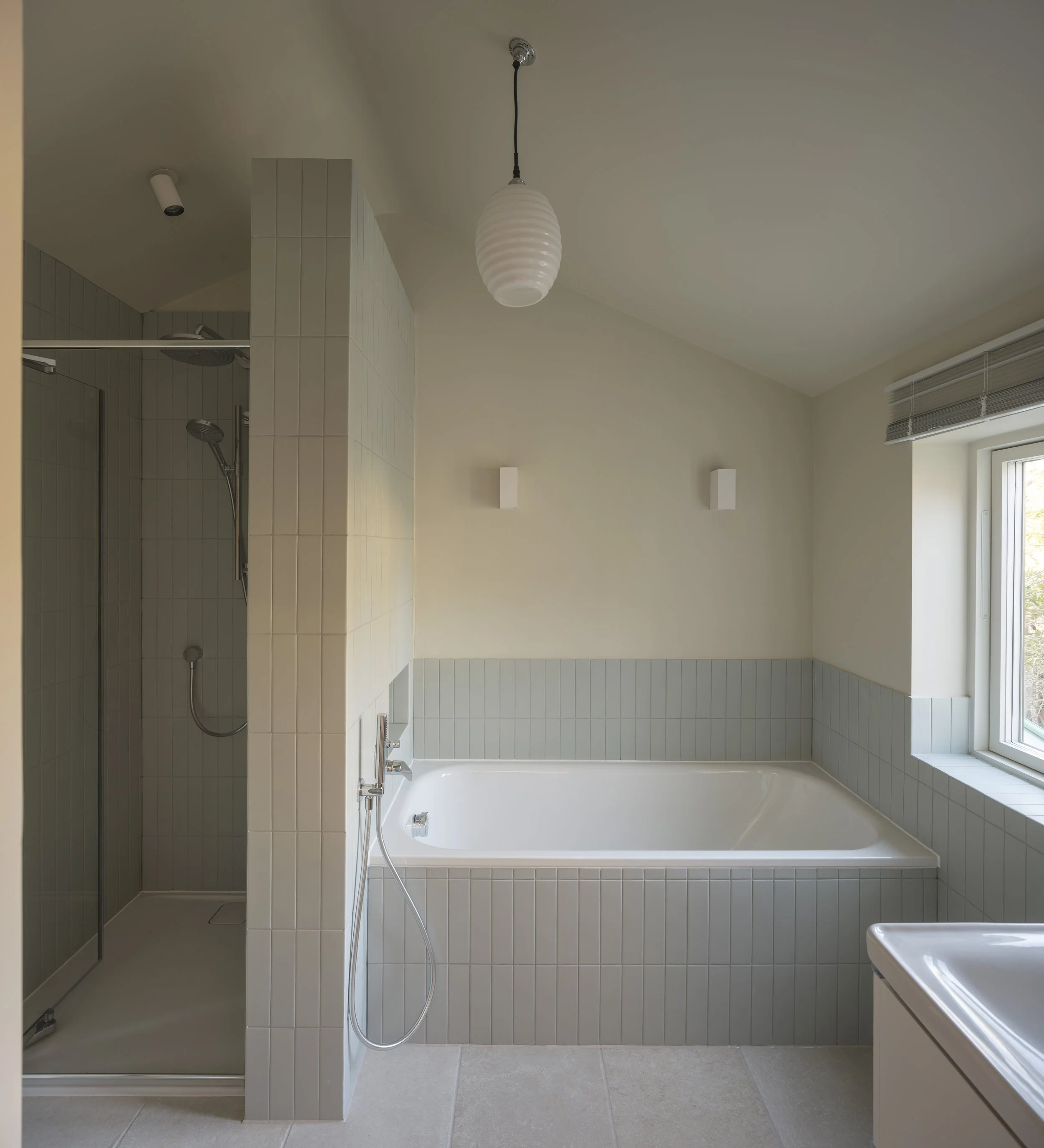 A modern bathroom with a shower area, bathtub, and vanity with sink, featuring a white and neutral color scheme, gray tiles, a window, and ceiling lights.