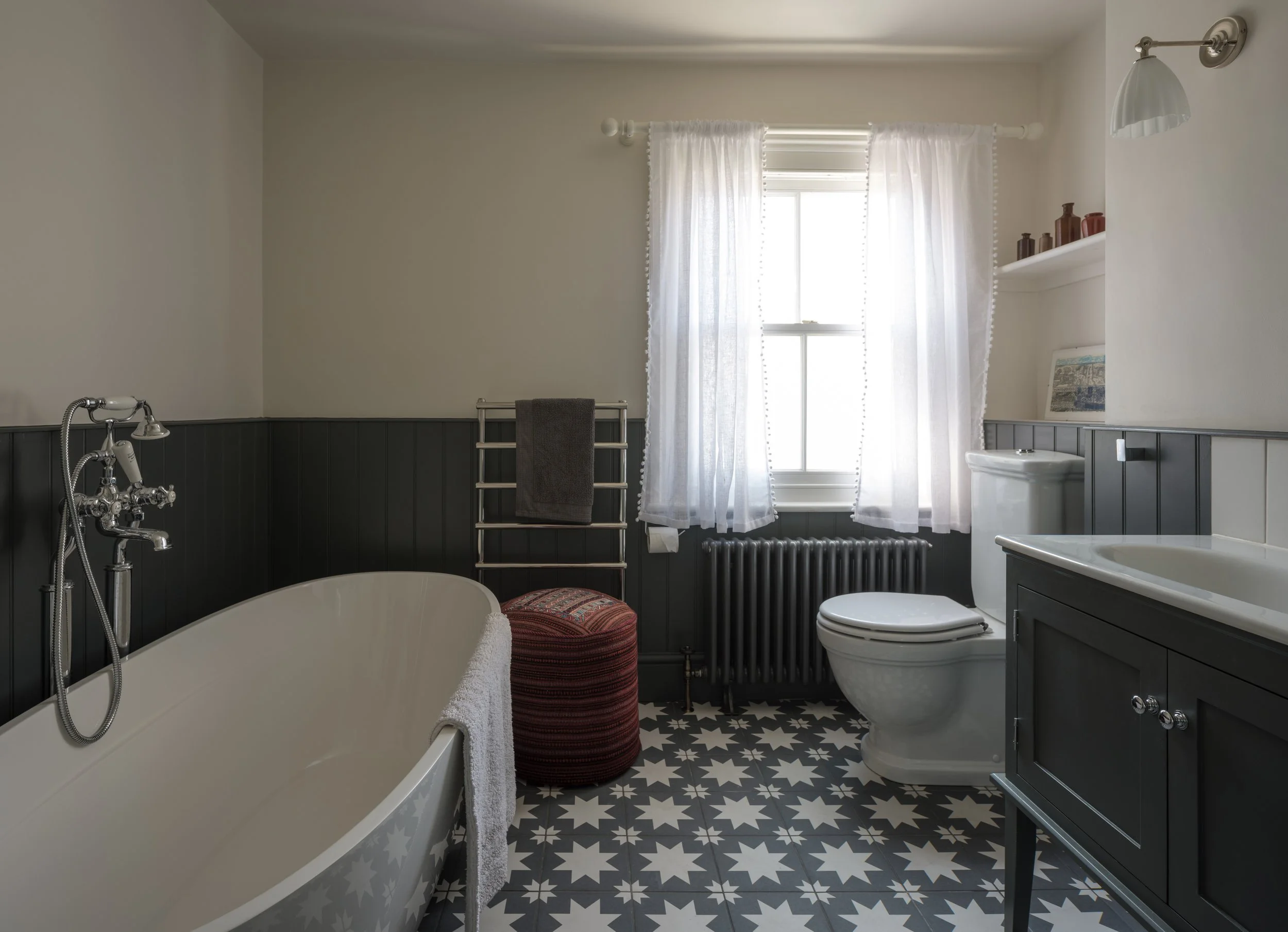 A bathroom with a white bathtub, black wainscoting, a window with white curtains, a toilet, a gray vanity, and patterned black and white floor tiles.