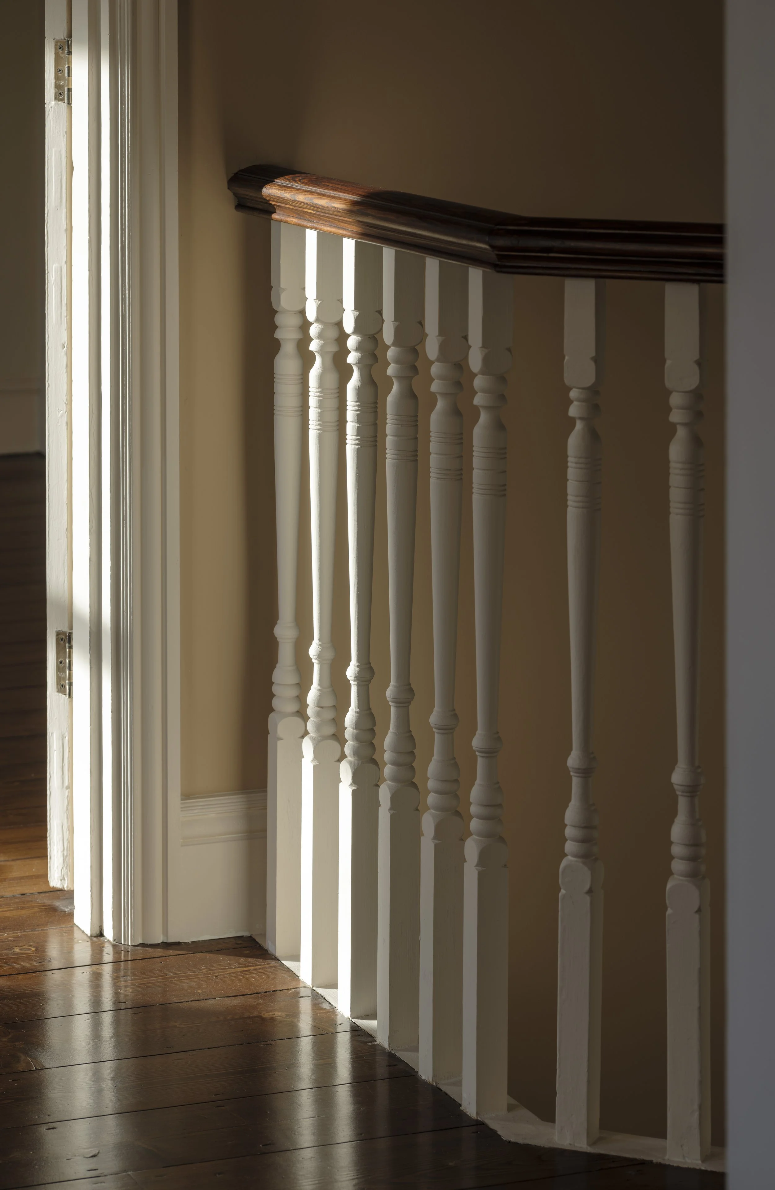 Interior view of a staircase with white balusters and a dark wooden handrail, with sunlight casting shadows on the wall.