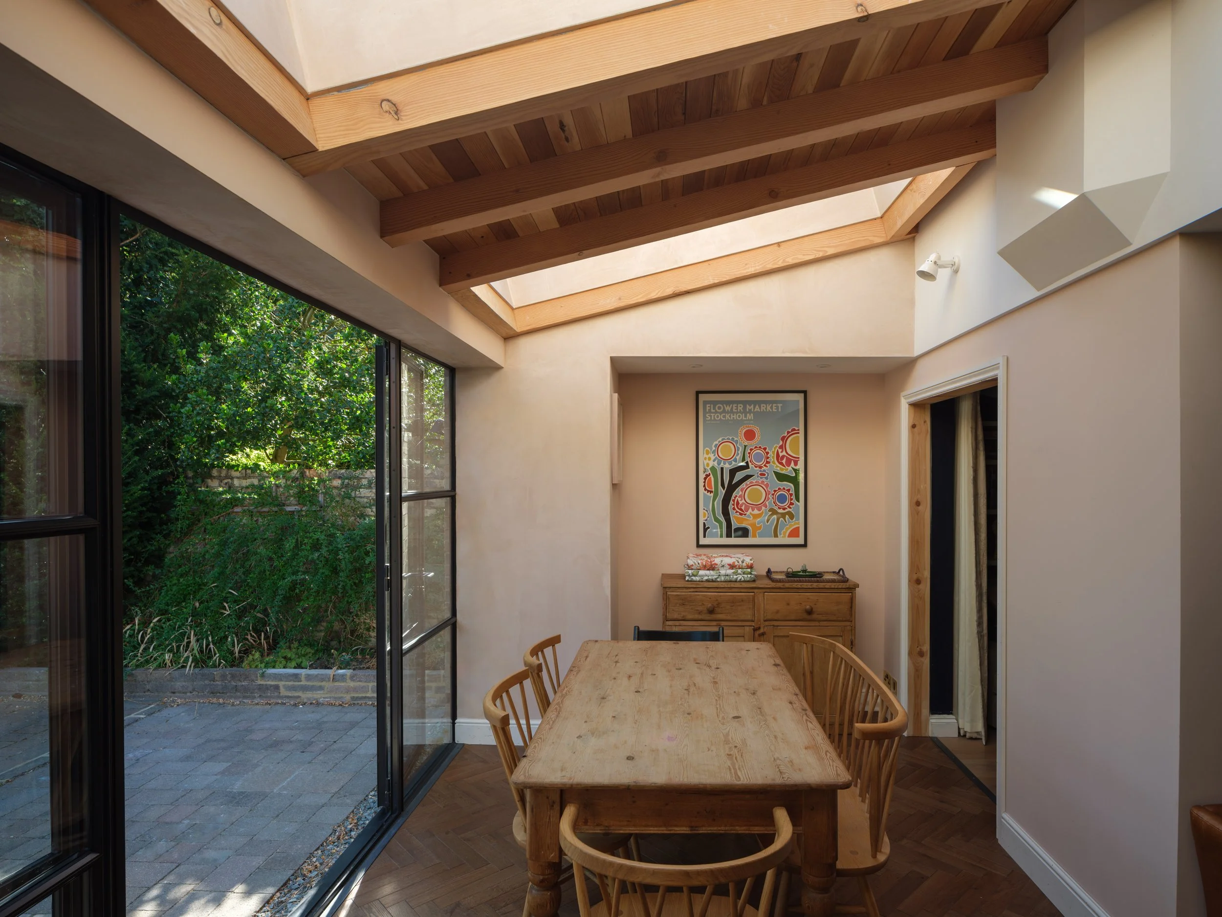 Interior of a dining area with a wooden table and chairs, large sliding glass doors opening to a garden, a framed colorful picture on the wall, and exposed wooden beams on the ceiling.