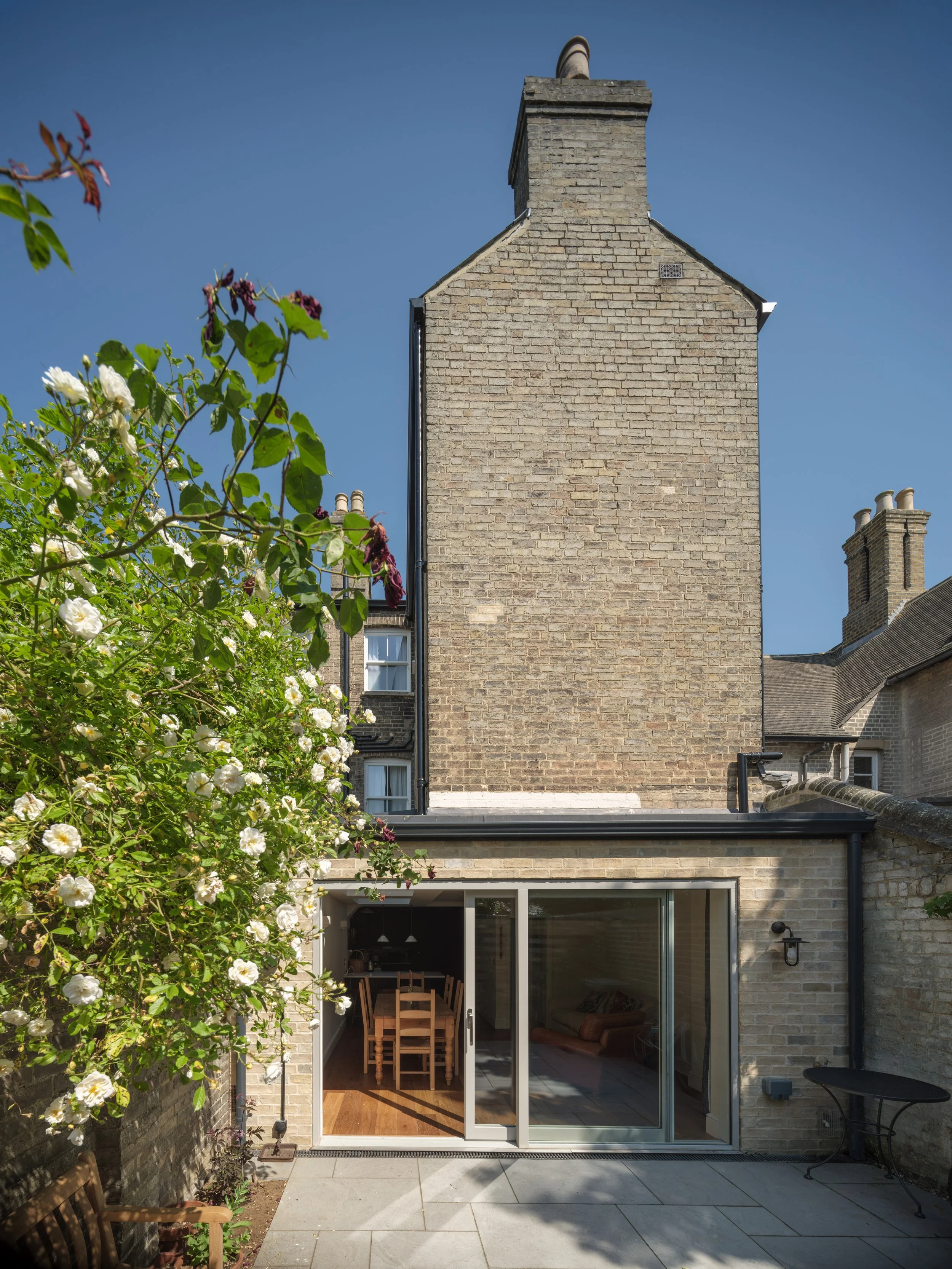 A backyard with a brick house, an open sliding glass door revealing a dining area with a wooden table and chairs, and a cozy living room with a couch inside. There are blooming white flowers and greenery on the left side, and a small black metal table on the right.
