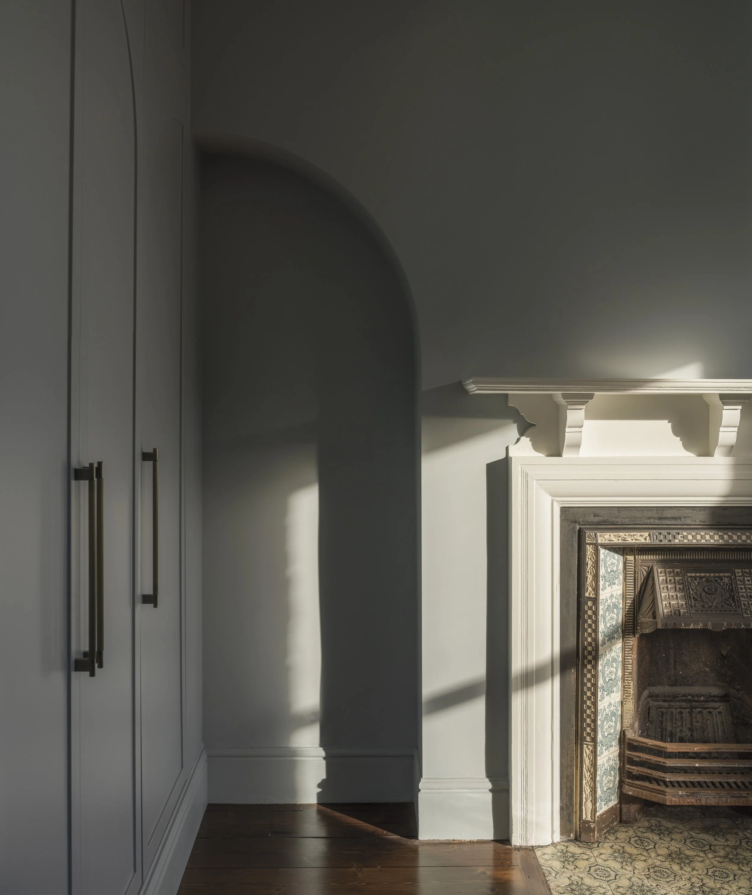 Interior view of a living room with a white fireplace, decorative molding, and a light-colored wall, illuminated by natural light from the right.