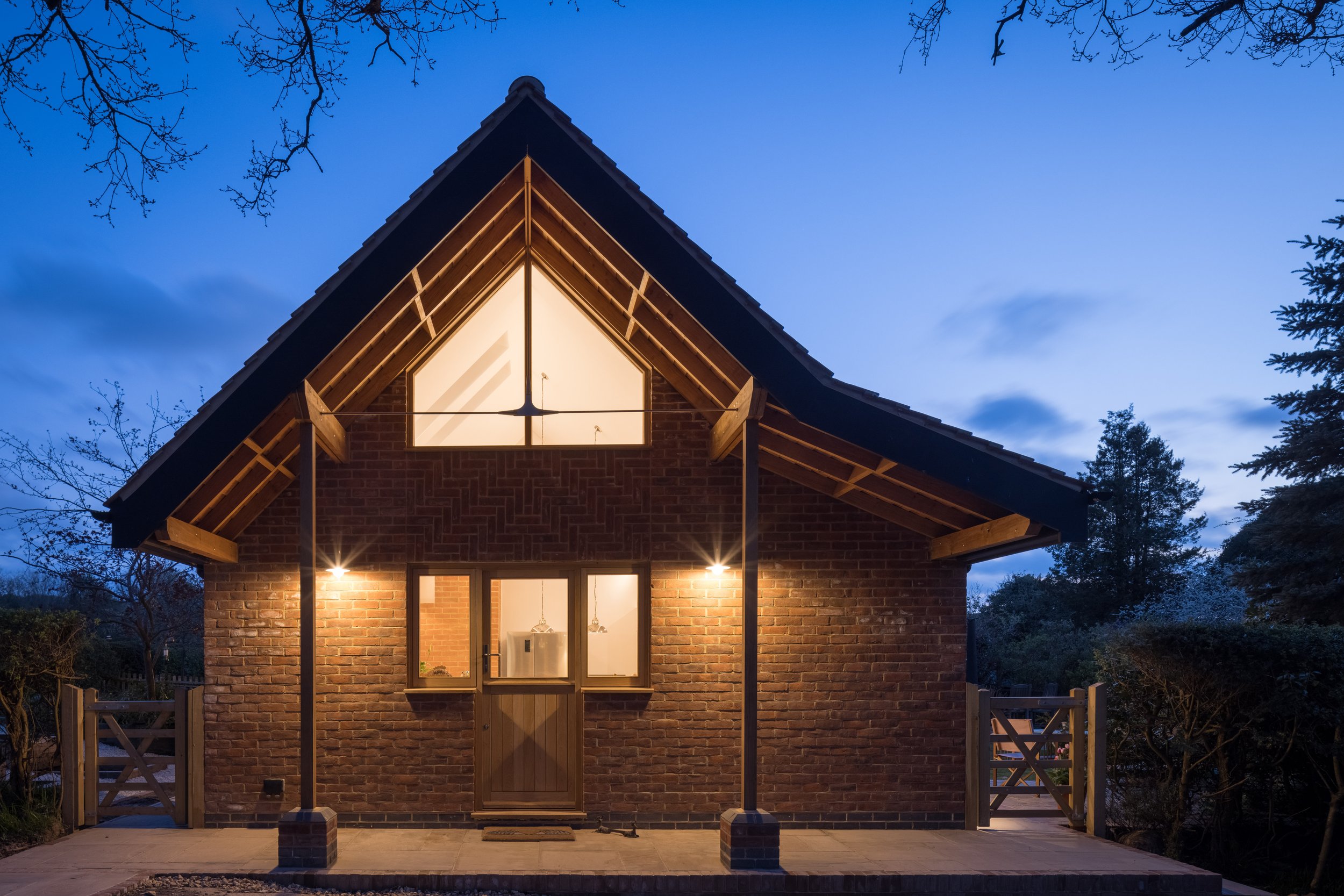 A brick house with a steep gabled roof, lit from inside, during dusk, surrounded by trees and a small porch with fencing.