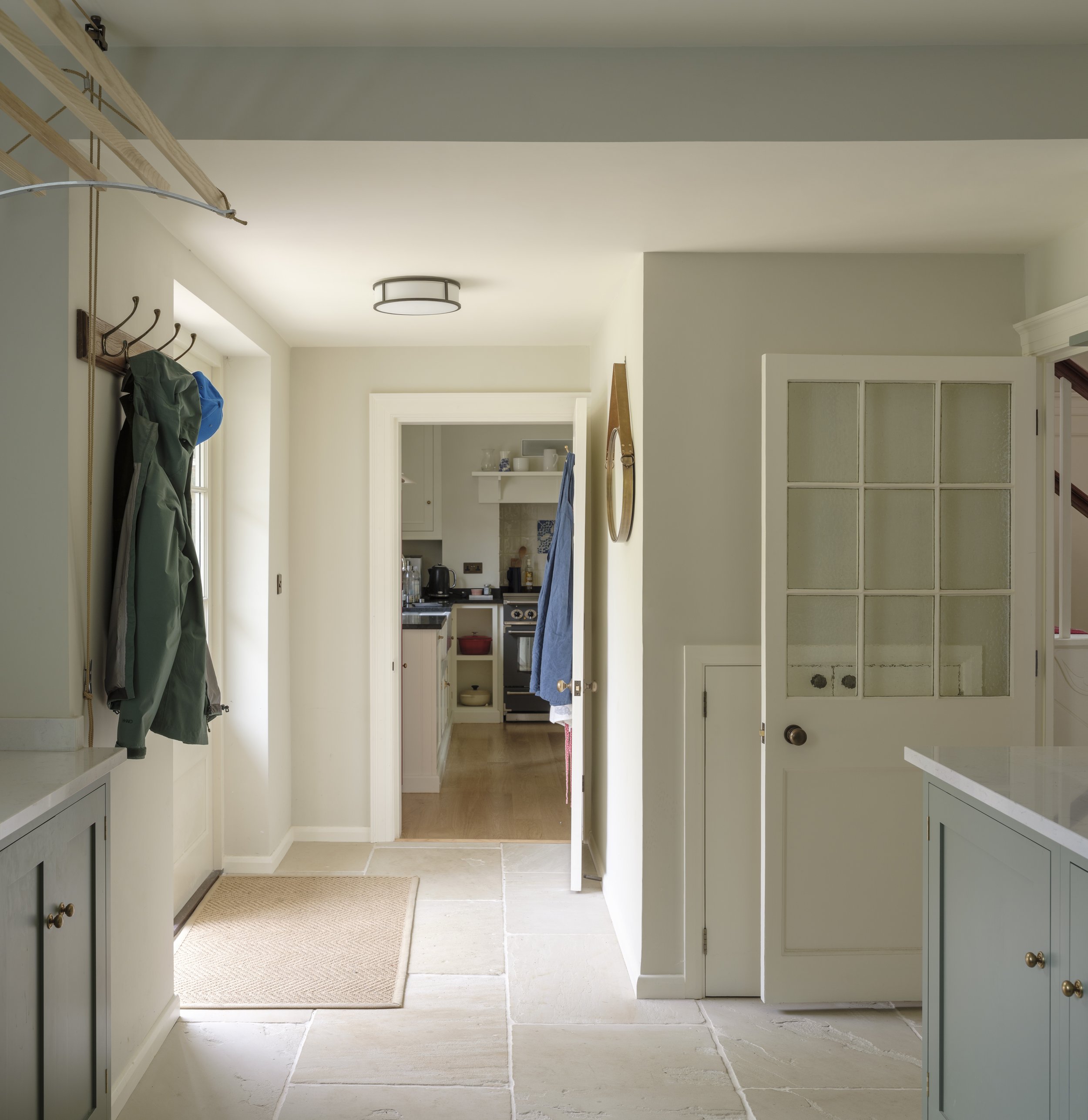 View of a kitchen and hallway with light-colored walls, wooden flooring, and a white door. Coat hooks with jackets hang on the wall. The kitchen has open shelving, a stove, and a small window. There is natural light coming in, and the ceiling has a m