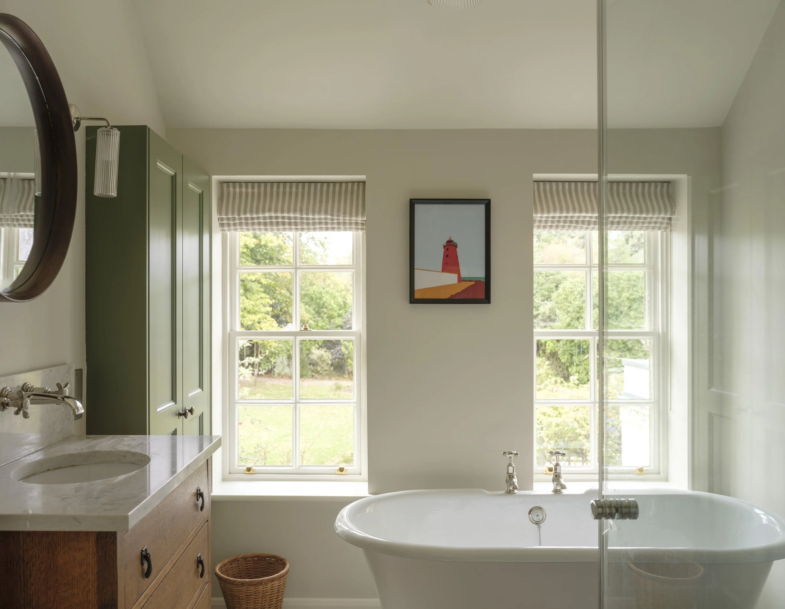 Bright bathroom with large window, garden view, framed lighthouse artwork, wooden vanity with marble countertop, round sink, woven basket, clawfoot bathtub, glass shower door, and striped window shades.