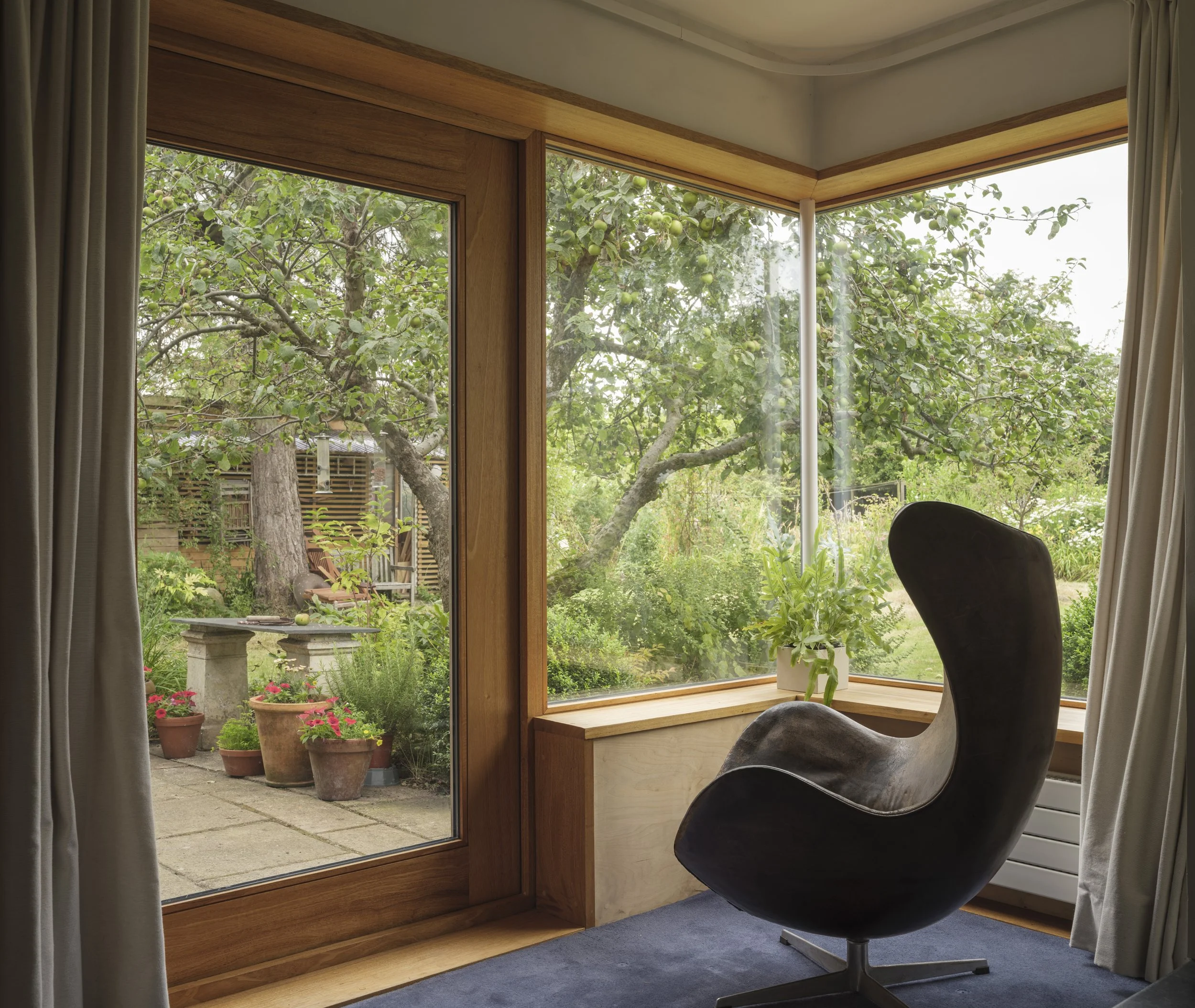 View of a garden with multiple potted plants outside a house, seen through glass sliding doors from inside a room. The room has curtains and a modern black chair near a wooden window seat.