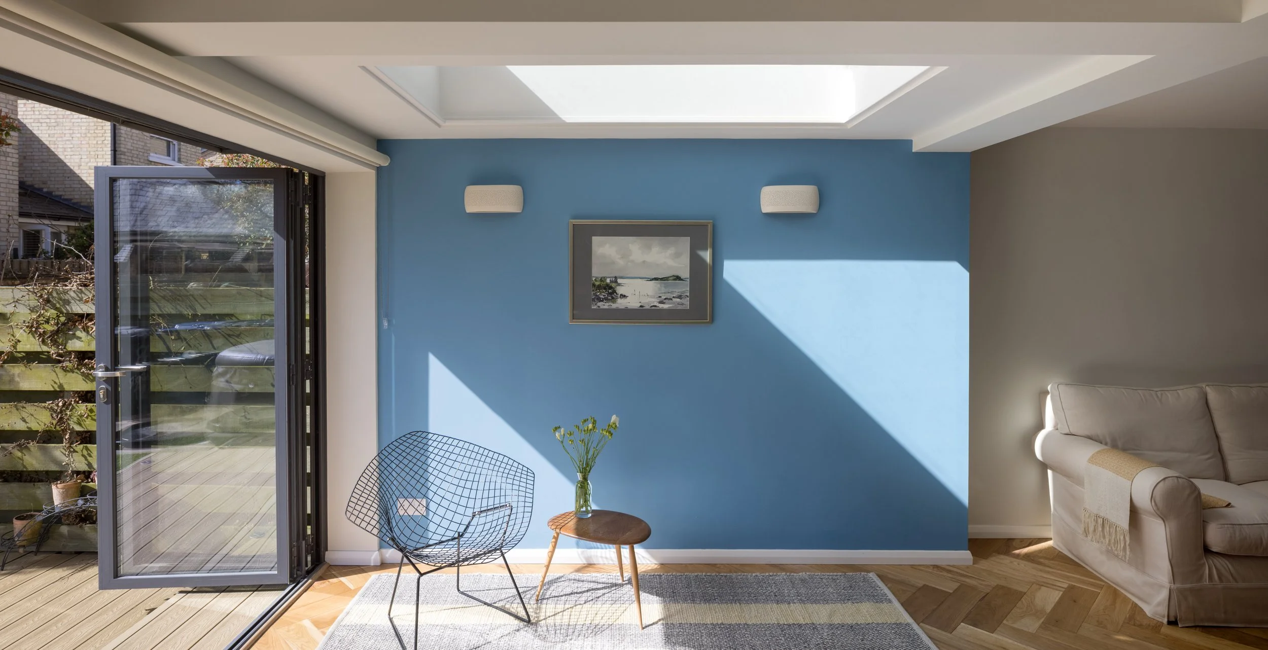 Sunlit living room with a blue accent wall, a framed picture of a seaside landscape, a beige sofa, a wire chair, a small wooden side table with a glass vase of yellow flowers, and a ceiling skylight. Outside view through a glass door shows a patio an