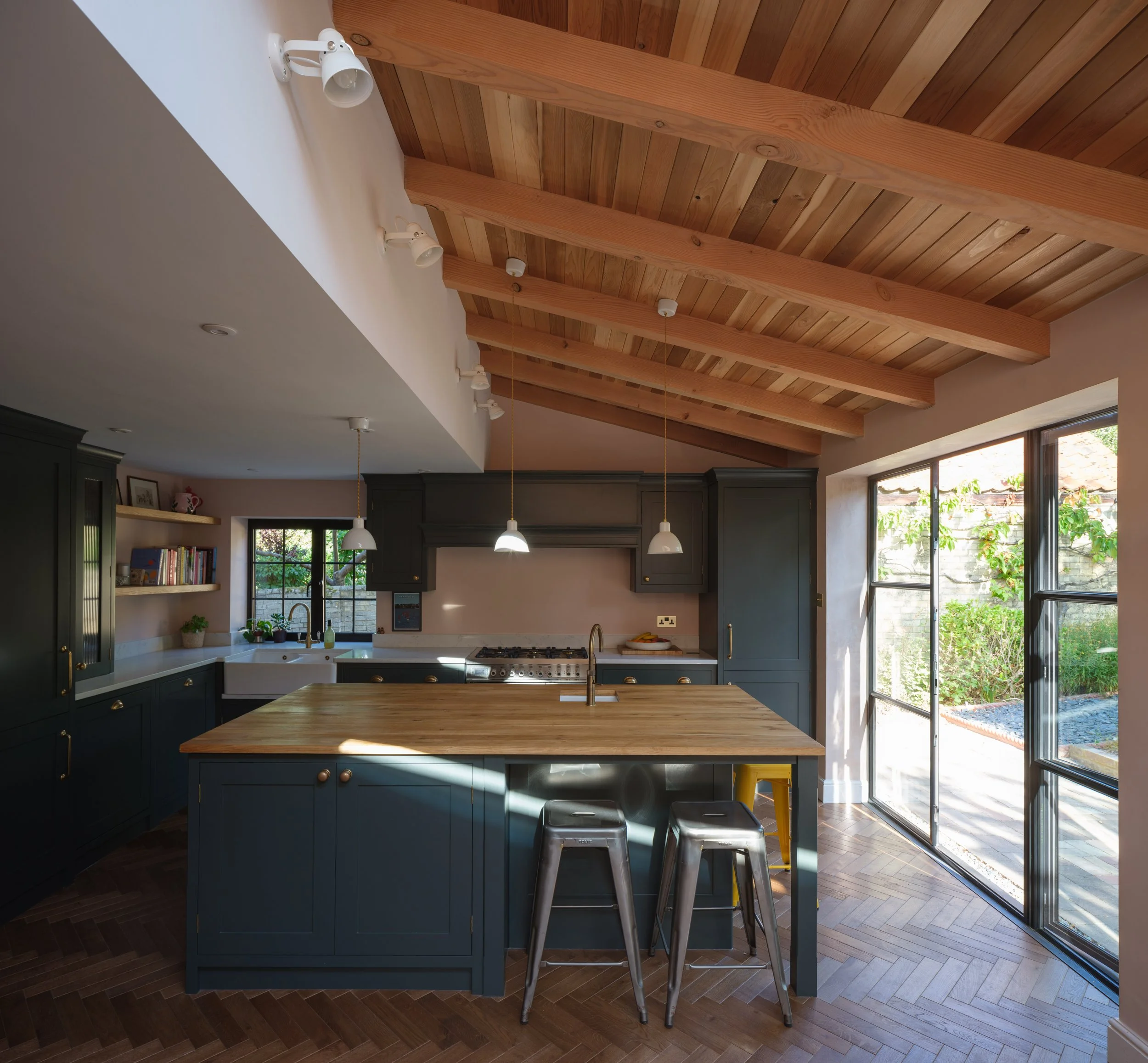 View of a modern kitchen with dark gray cabinets, a wooden kitchen island, stainless steel stools, and large windows bringing in natural light, with a wooden ceiling featuring exposed beams.