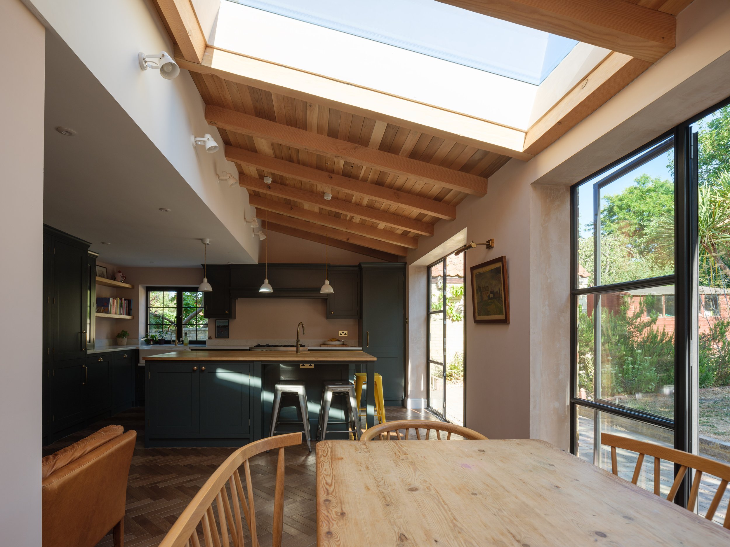 A kitchen with wooden ceiling beams and a large skylight, dark cabinets, open shelves with books, pendant lights, and large windows showing green trees outside.