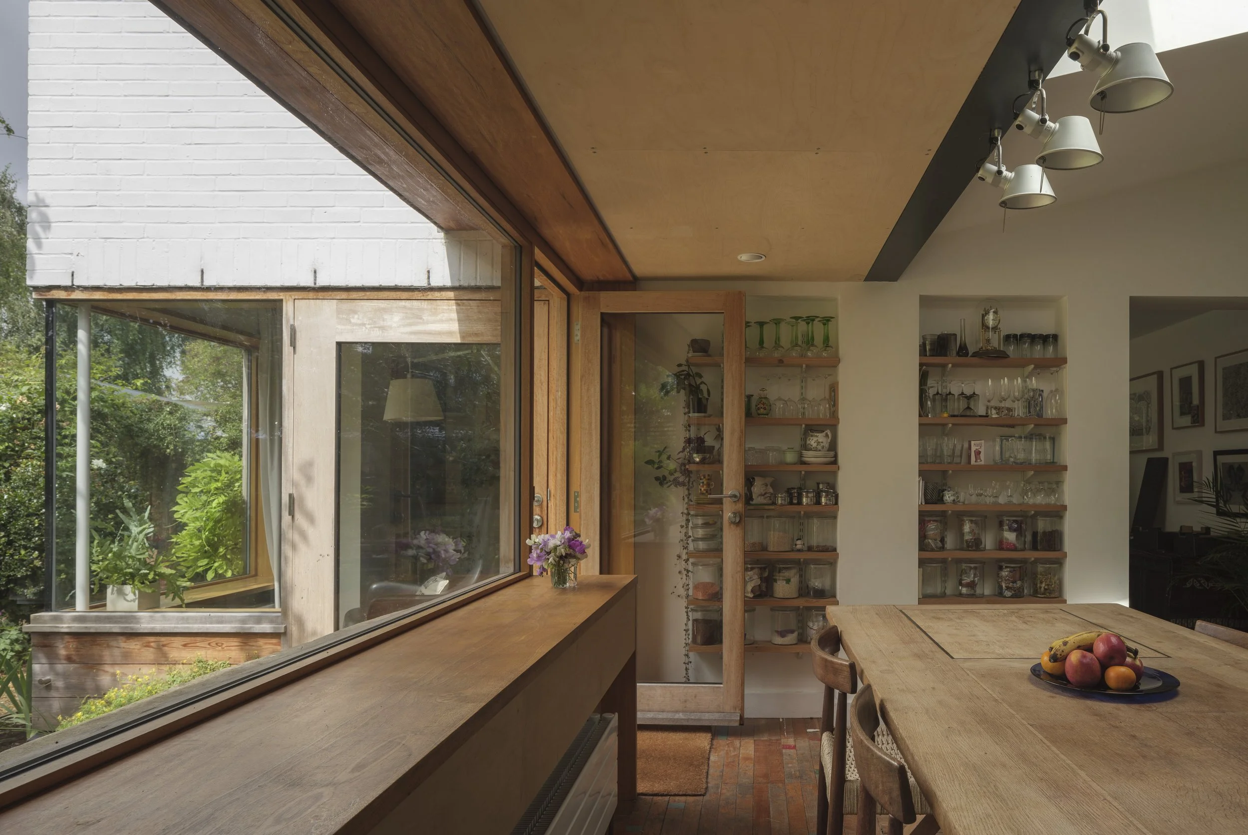A view of a cozy kitchen with large windows overlooking a garden, a wooden dining table with apples, and open shelves with glassware and plants.