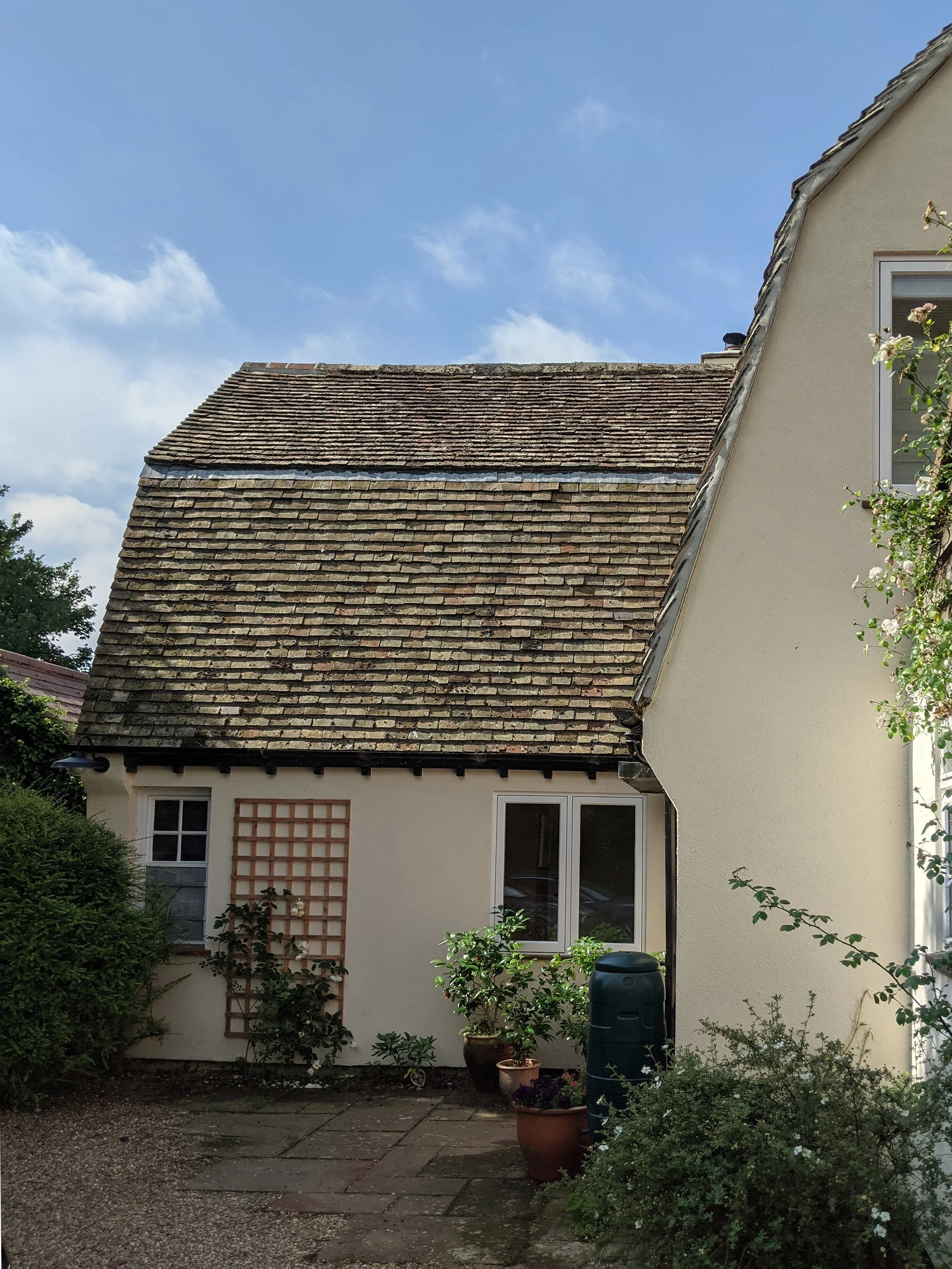 A garden area outside a house, with potted plants, bushes, and a water barrel, next to a white house with large windows and a trellis. The house has a tiled roof, and the sky is partly cloudy.
