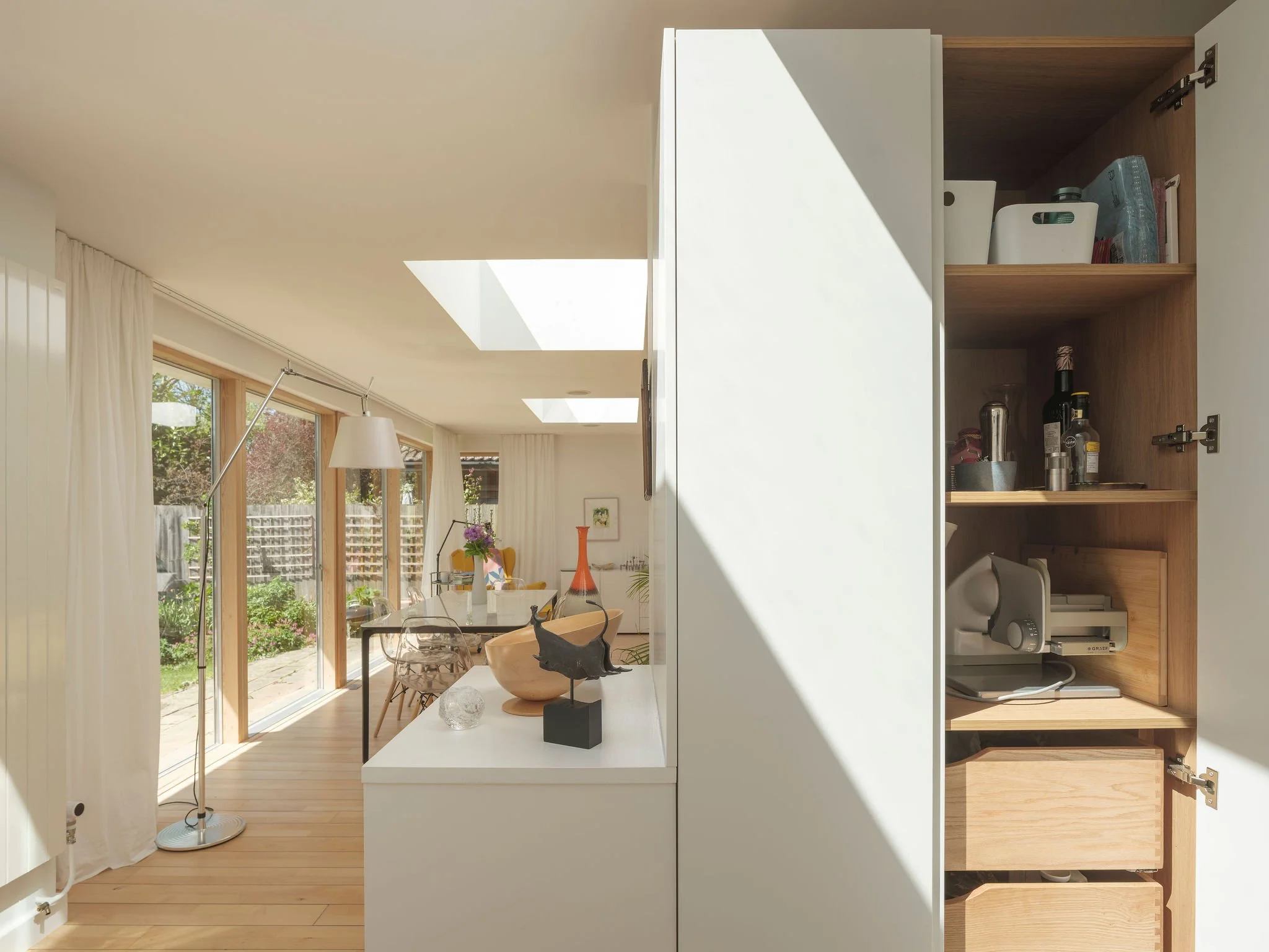 Open kitchen cabinet with shelves containing bottles, jars, and kitchen items, and a view into a bright dining area with large windows and curtains.
