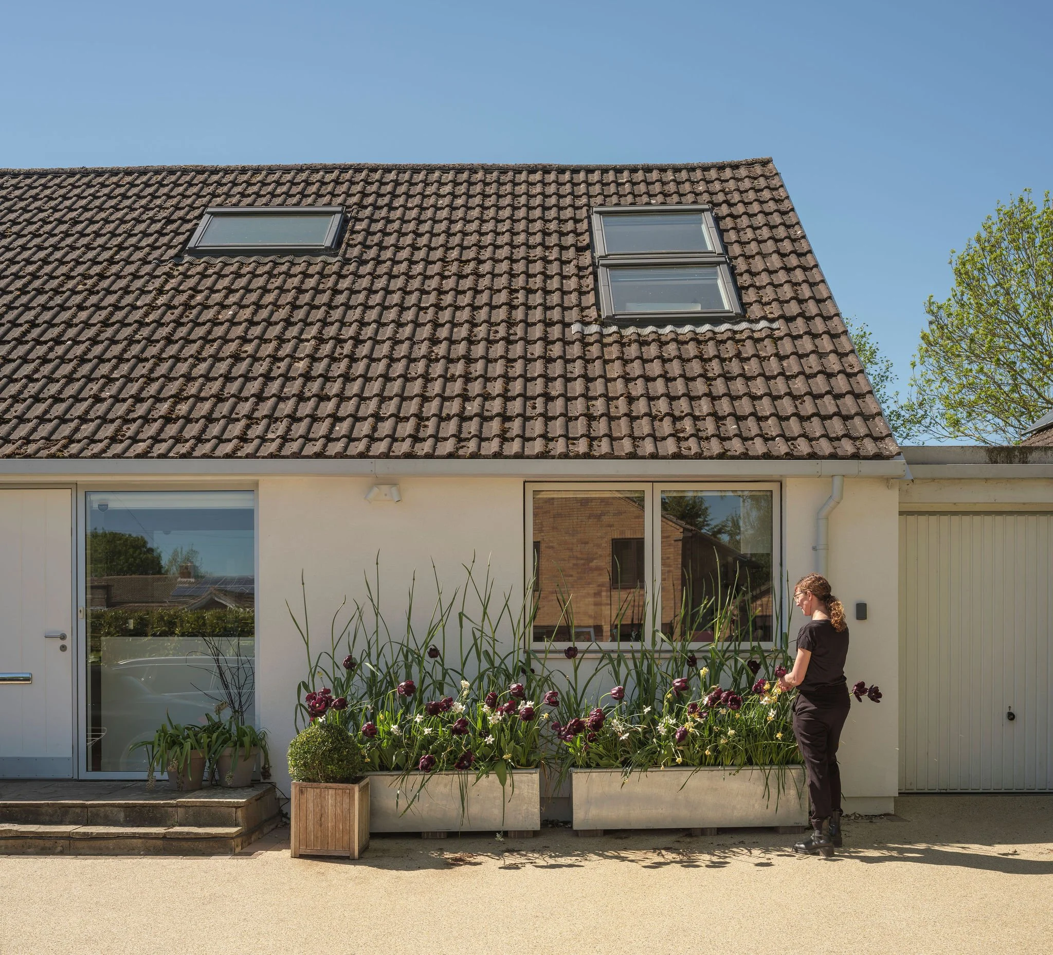 A woman standing outside in front of a house with a sloped roof and three windows, next to a flower bed with purple and white flowers, and a tree with green leaves on the right side. The house has a white exterior and large glass sliding doors.