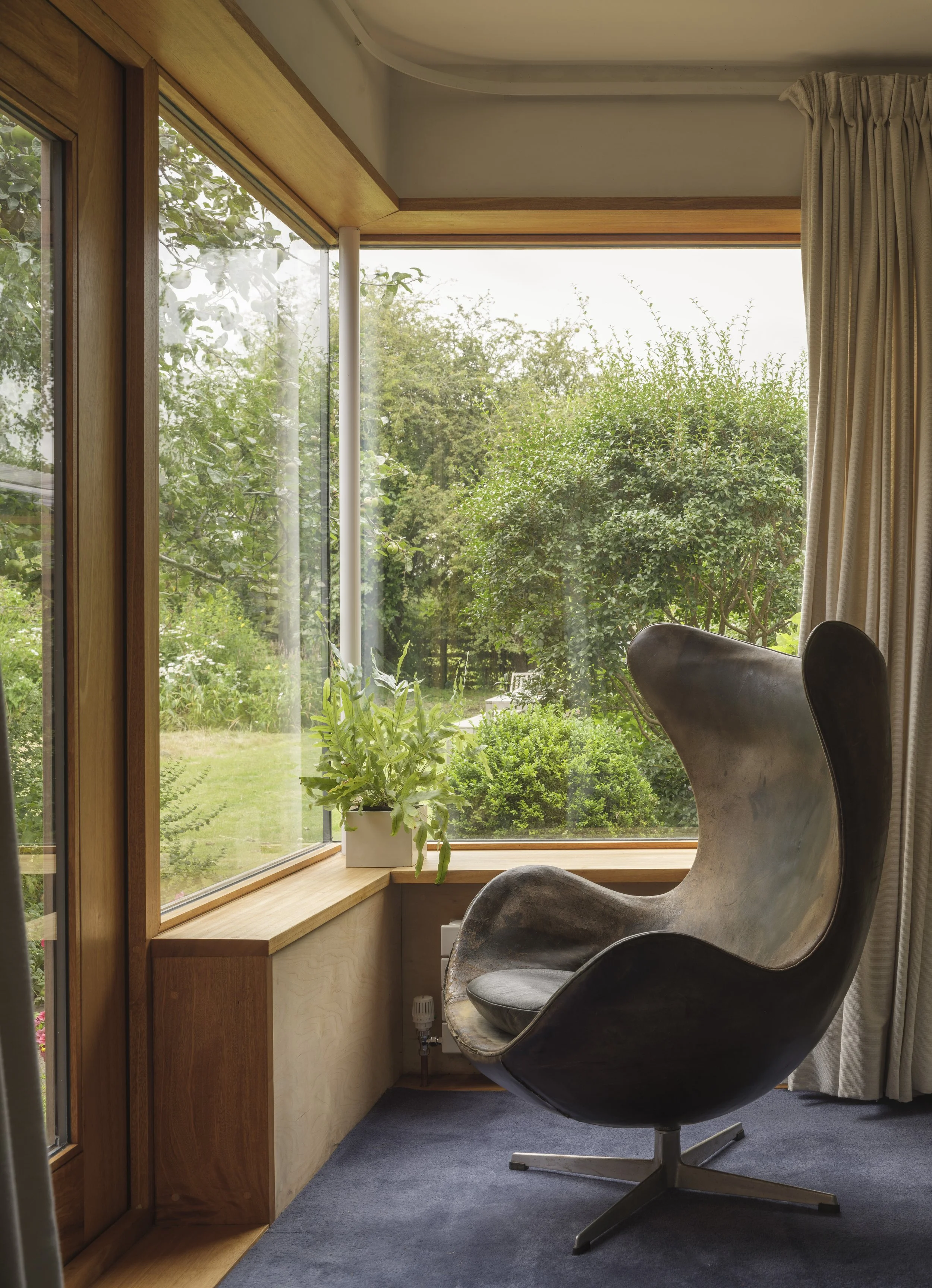 A modern black leather chair positioned by a large window with a view of green trees, a potted plant on the window sill, beige curtains to the right, and a blue carpeted floor.