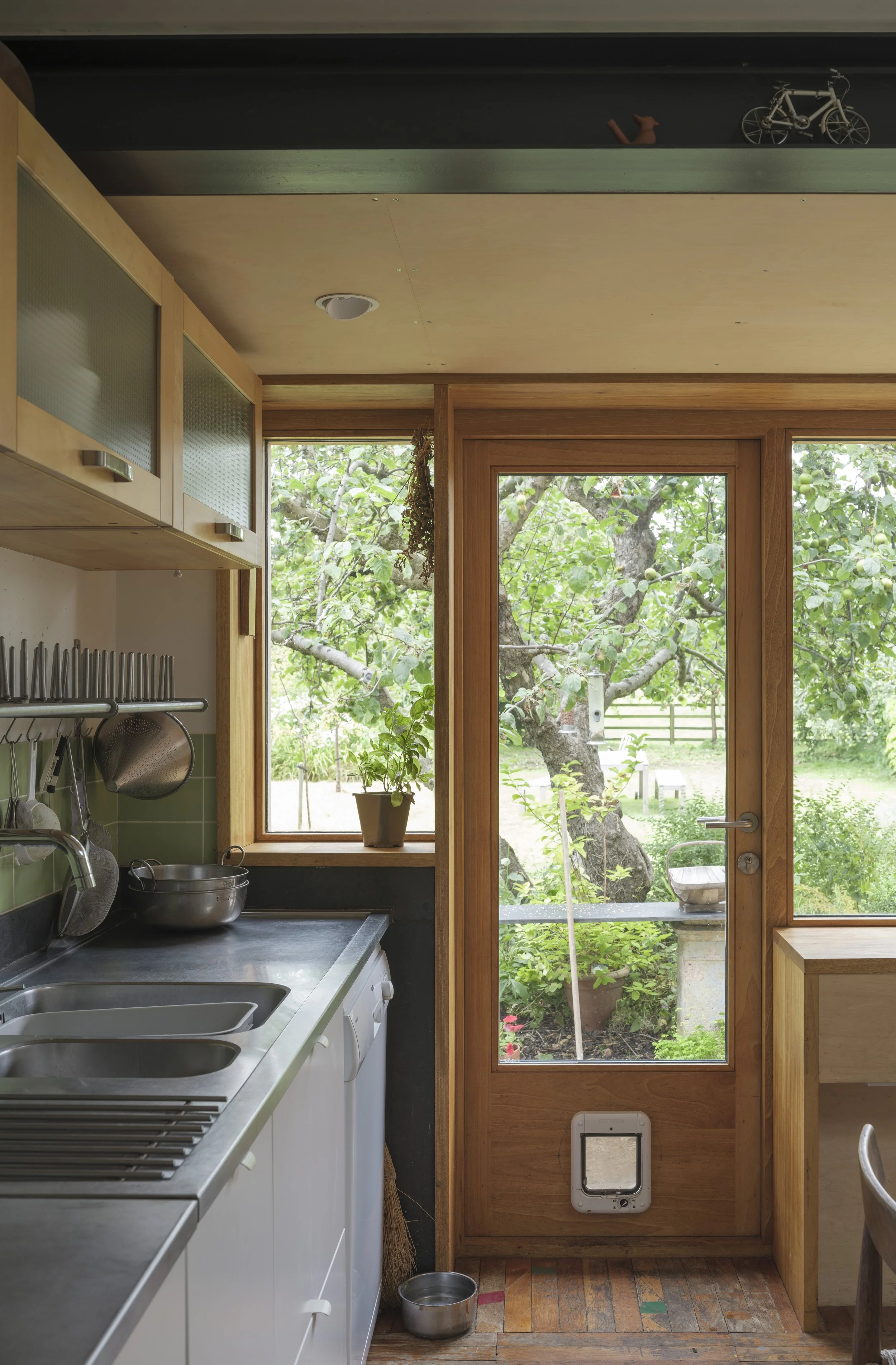 View of a kitchen with a window and door overlooking a garden with trees, a potted plant on the windowsill, and kitchen utensils hanging on the wall.