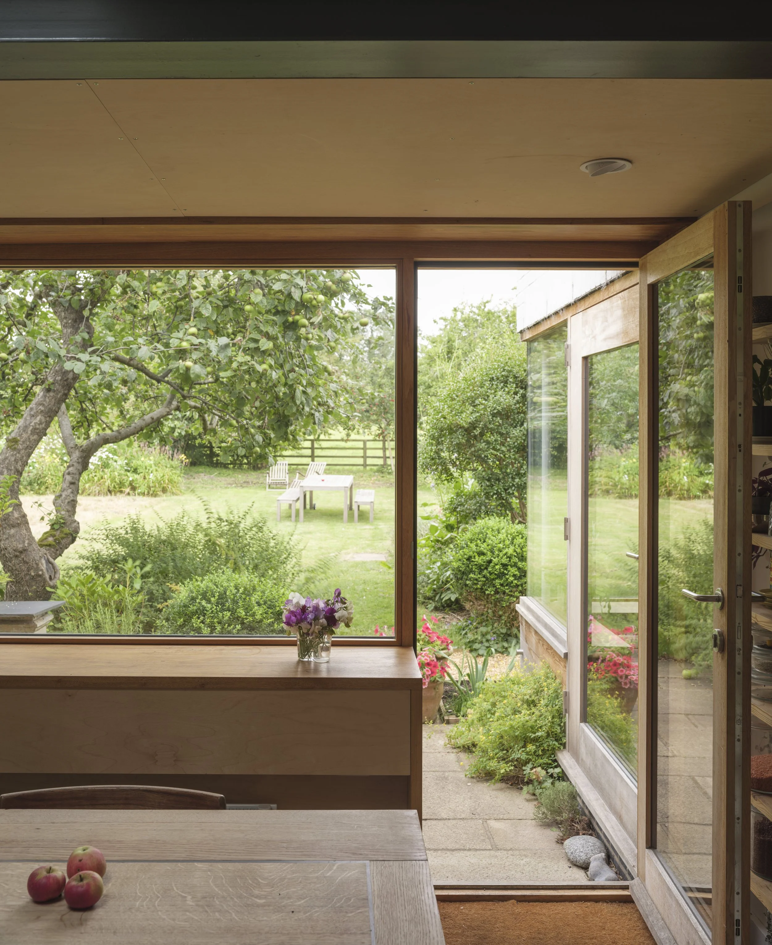 View from inside a house through glass windows and a sliding door, showing a lush green garden with a tree bearing green apples, a wooden bench, and a white table outdoors, with potted plants and pink flowers near the entrance.
