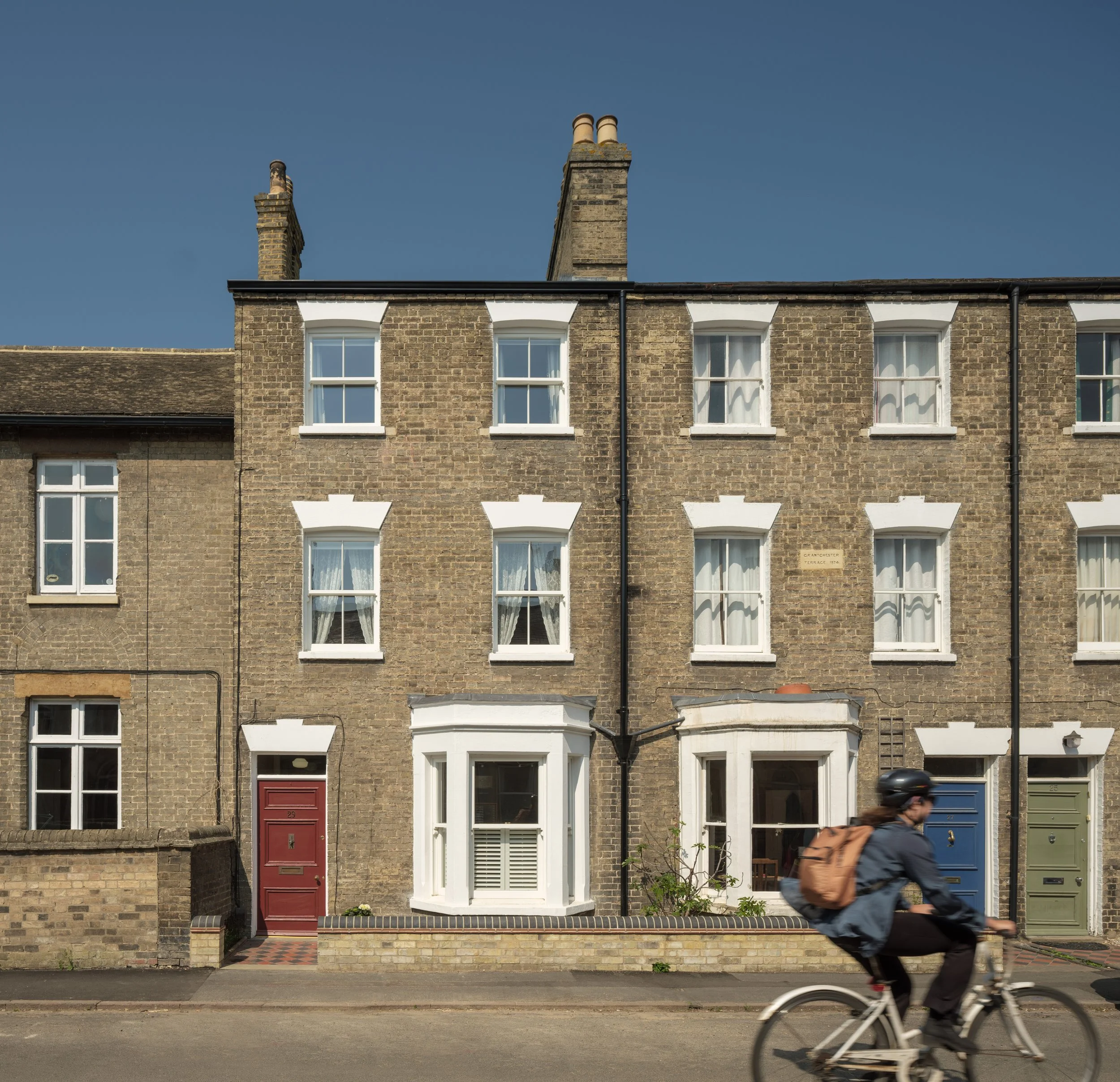 Front view of a brick residential building with multiple windows and two chimneys, one with double pipes, against a clear blue sky. A person with a backpack is riding a bicycle on the street in the foreground.