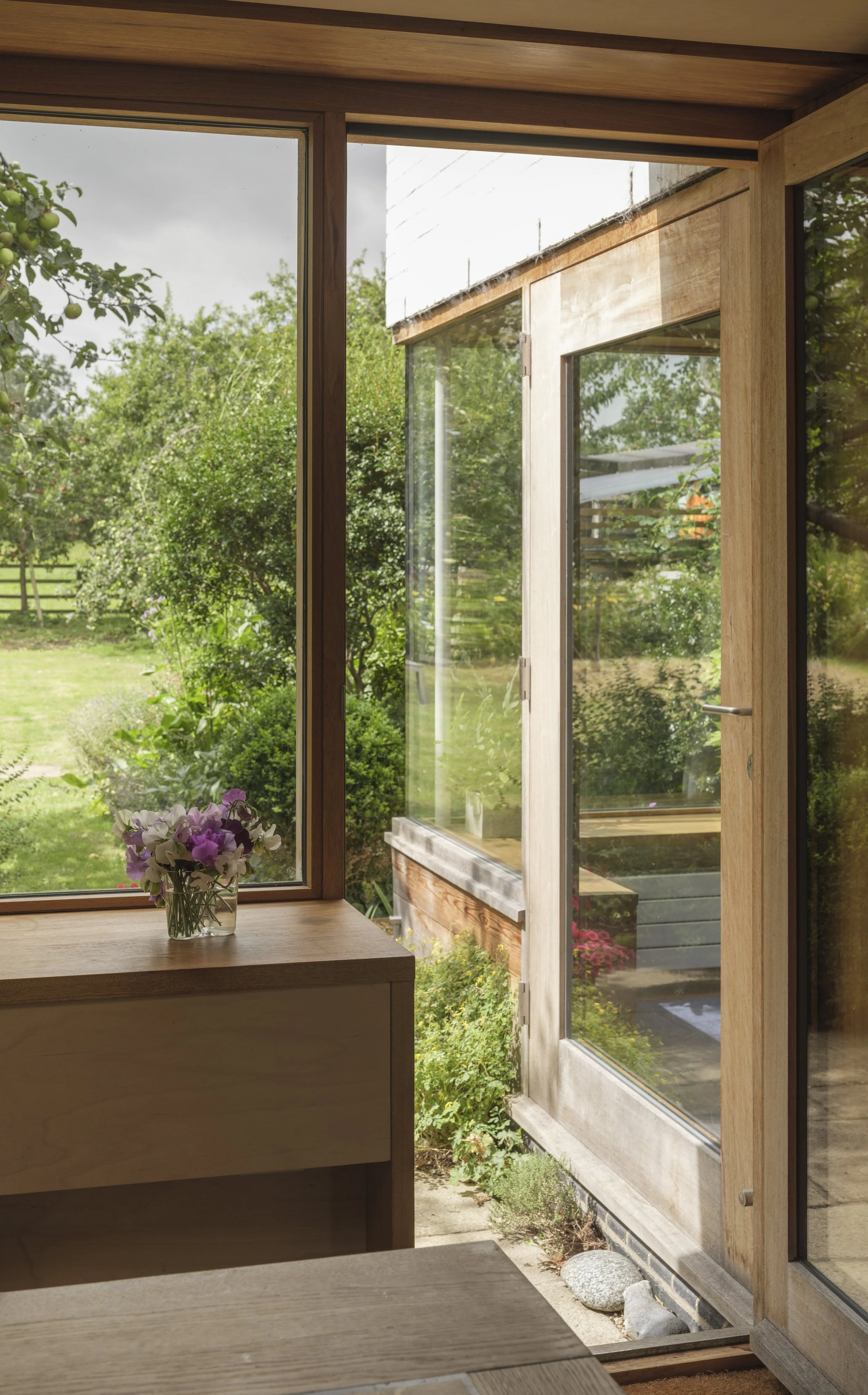 View from inside a house looking out through glass doors to a lush green garden with trees and bushes, blue sky with some clouds, and a wooden-structure outdoor space.