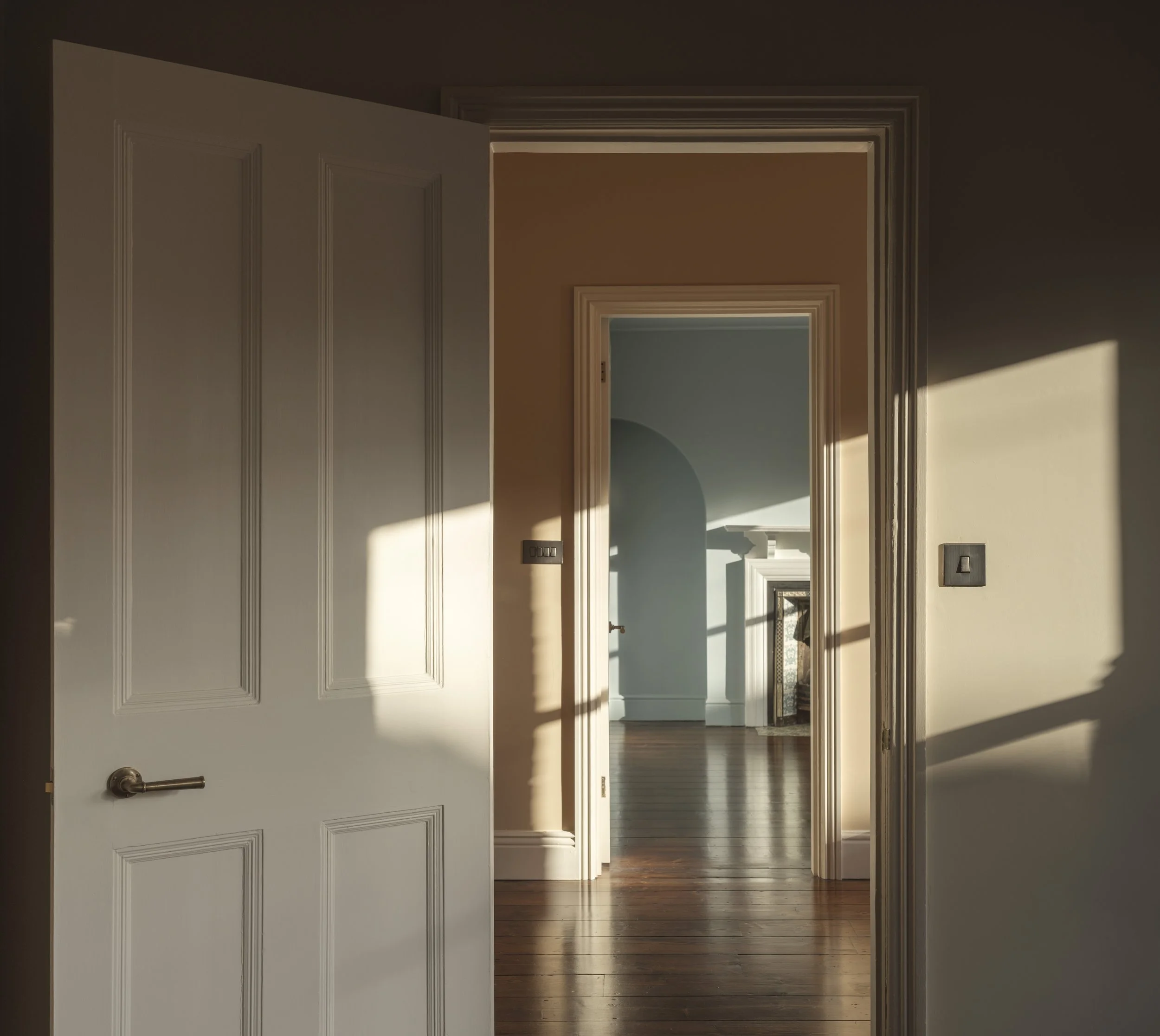Open white door leading into a hallway with wooden flooring and multiple doorways visible, sunlight casting shadows on the floor.