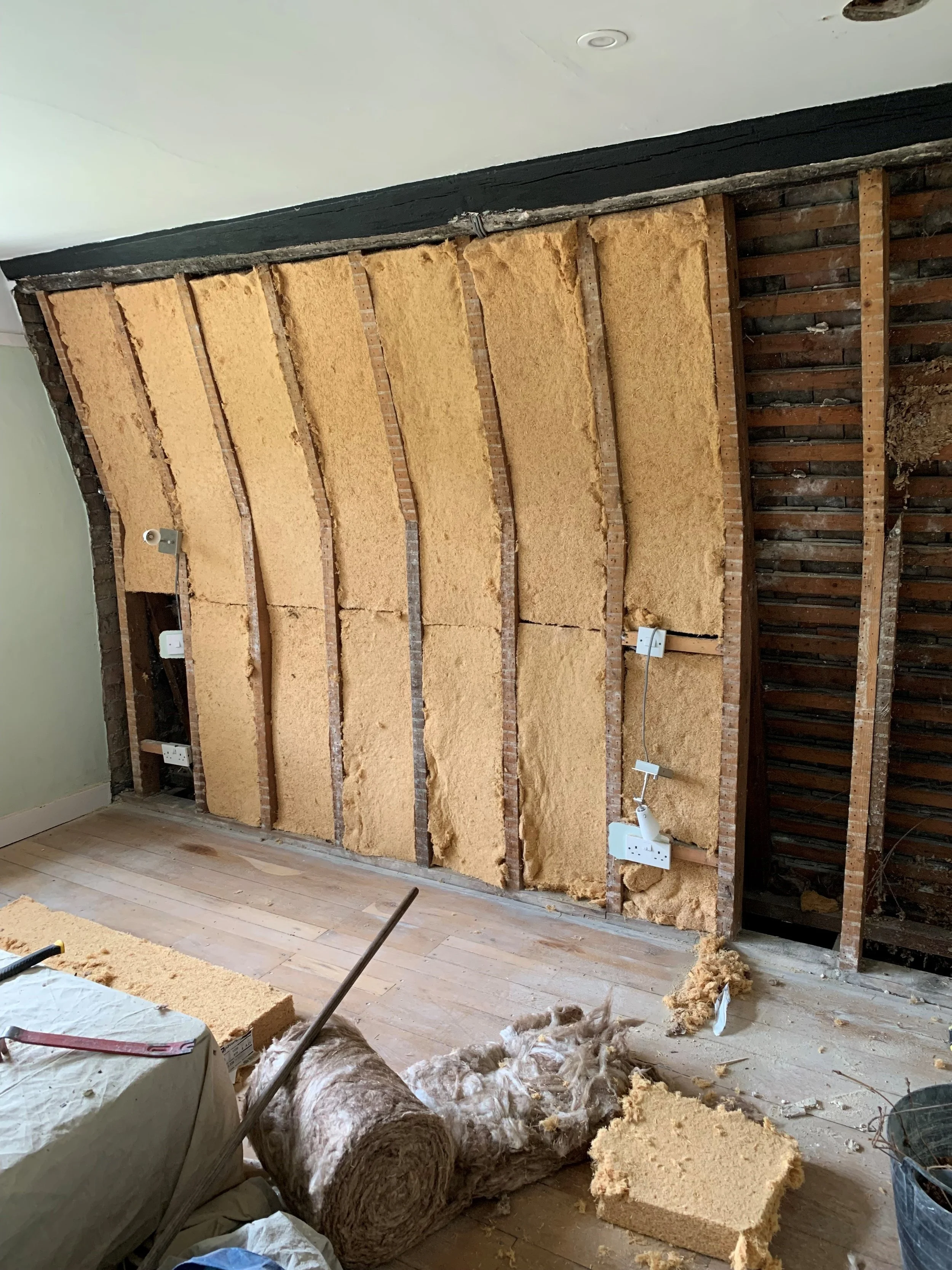 A partially renovated room with exposed wall studs, insulation being installed, and various construction tools and materials on the floor, including rolls of insulation and a large bag with insulation.