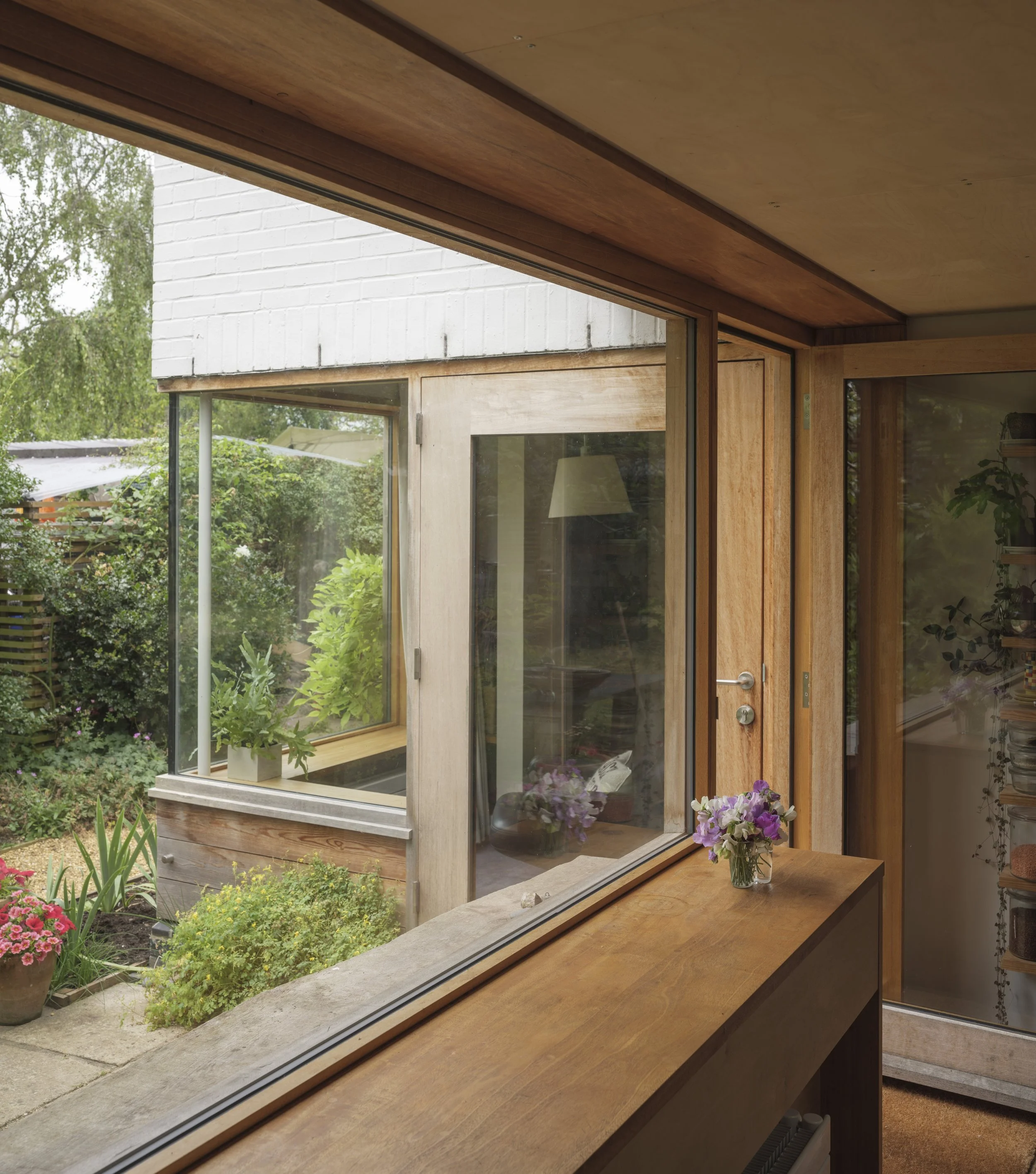 A window with a wooden frame, overlooking a garden with green plants and flowers. Inside, there is a wooden table with a vase of purple and white flowers.