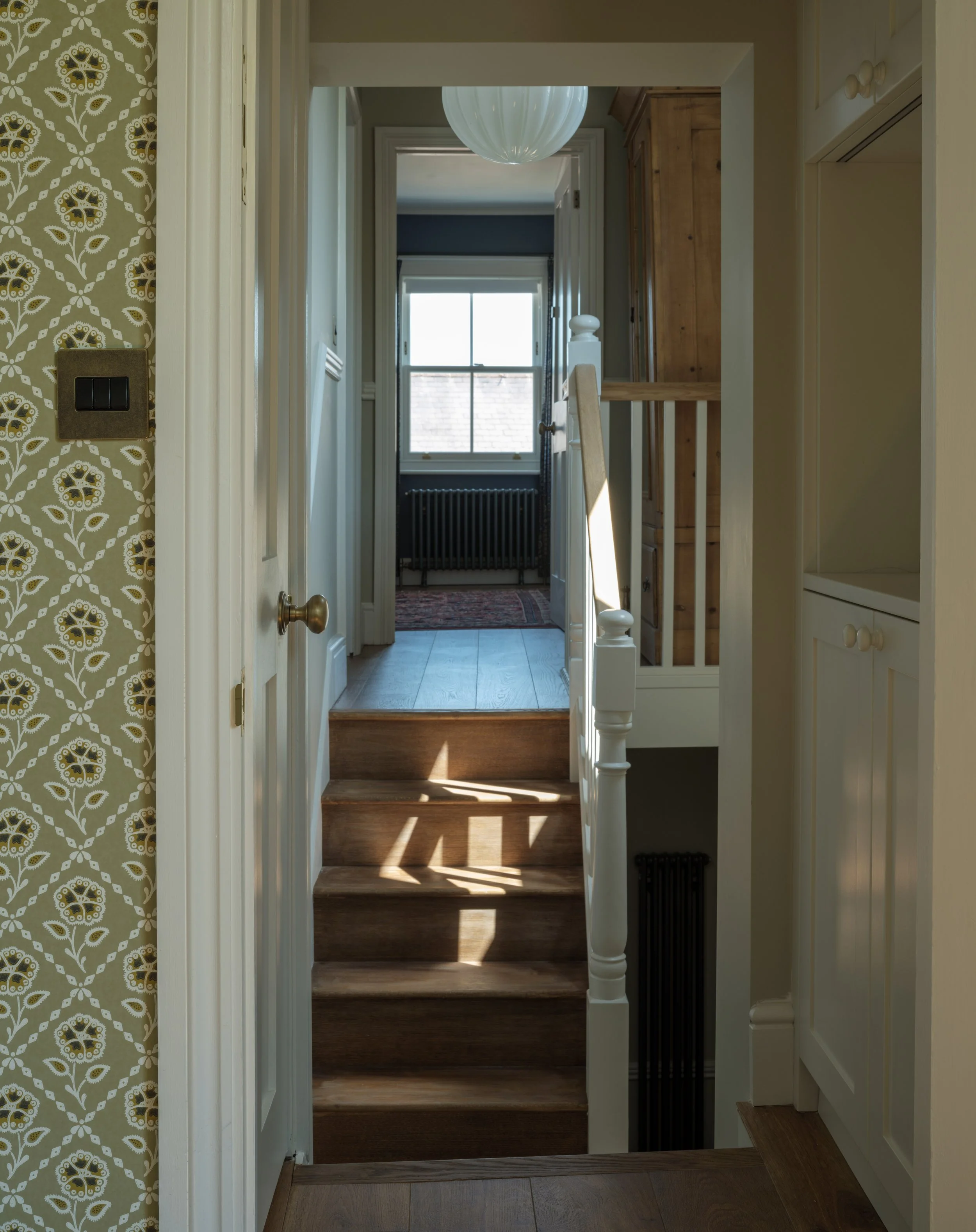 View of a hallway leading to a window with sunlight, a wooden staircase, a patterned wallpaper on the left, and white cabinets on the right.