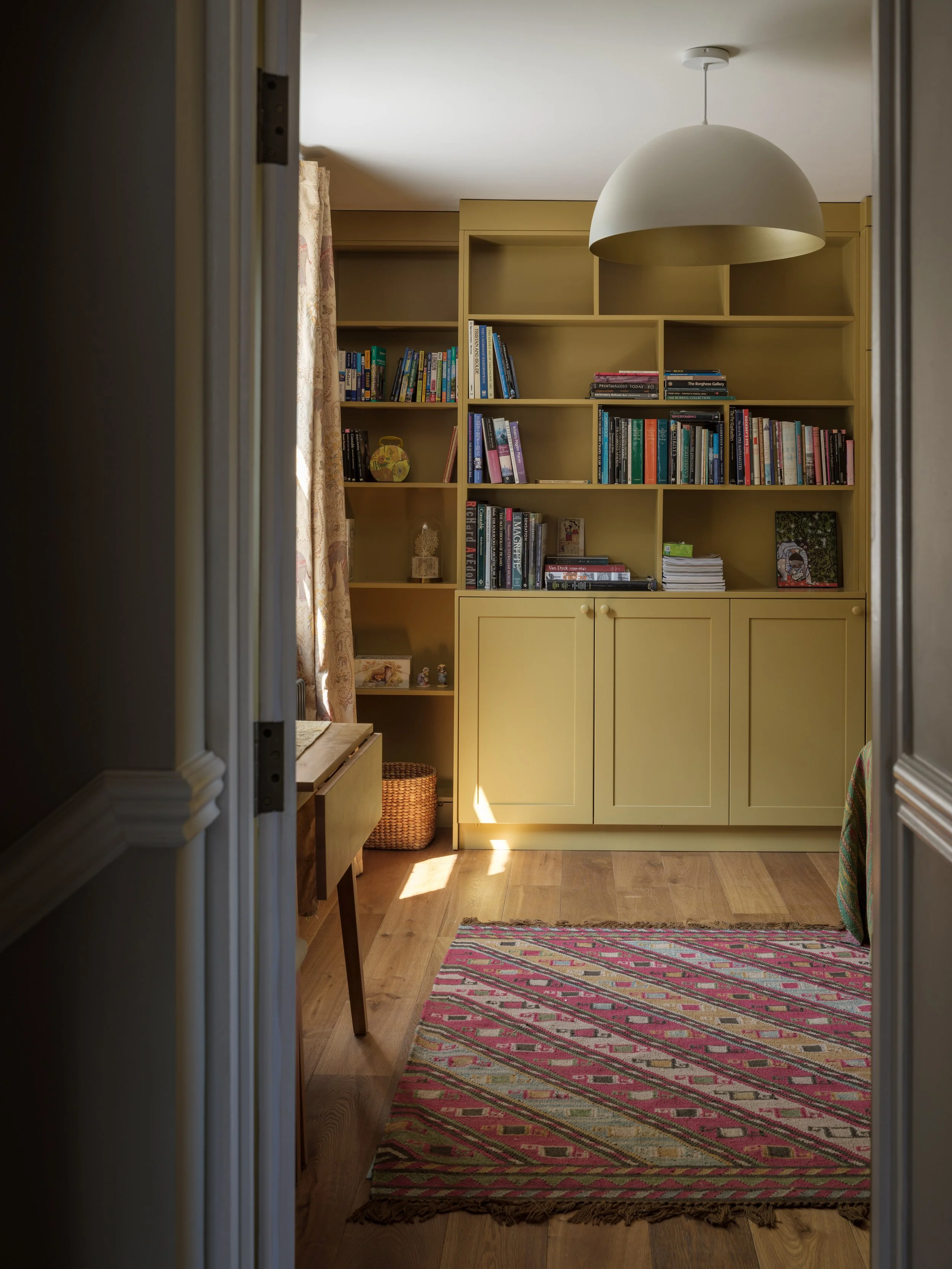 A view into a room with built-in yellow shelving, a multicolored rug, a wooden bench, and a colorful tapestry hanging on the side. There is a large white ceiling light and a window with floral curtains on the left side.