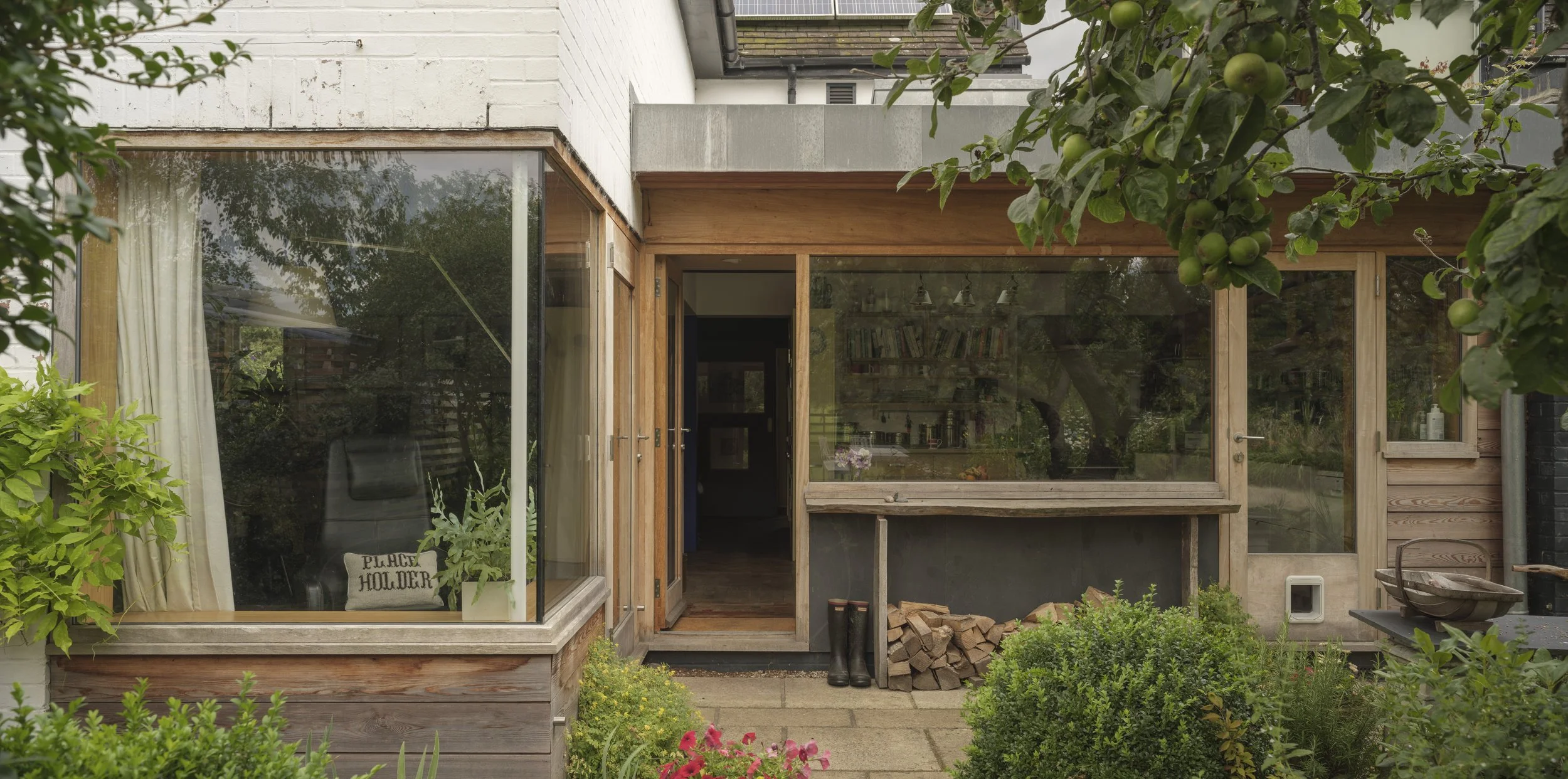 A backyard view of a house with large glass windows and wooden framing, surrounded by lush greenery and a small garden with colorful flowers.
