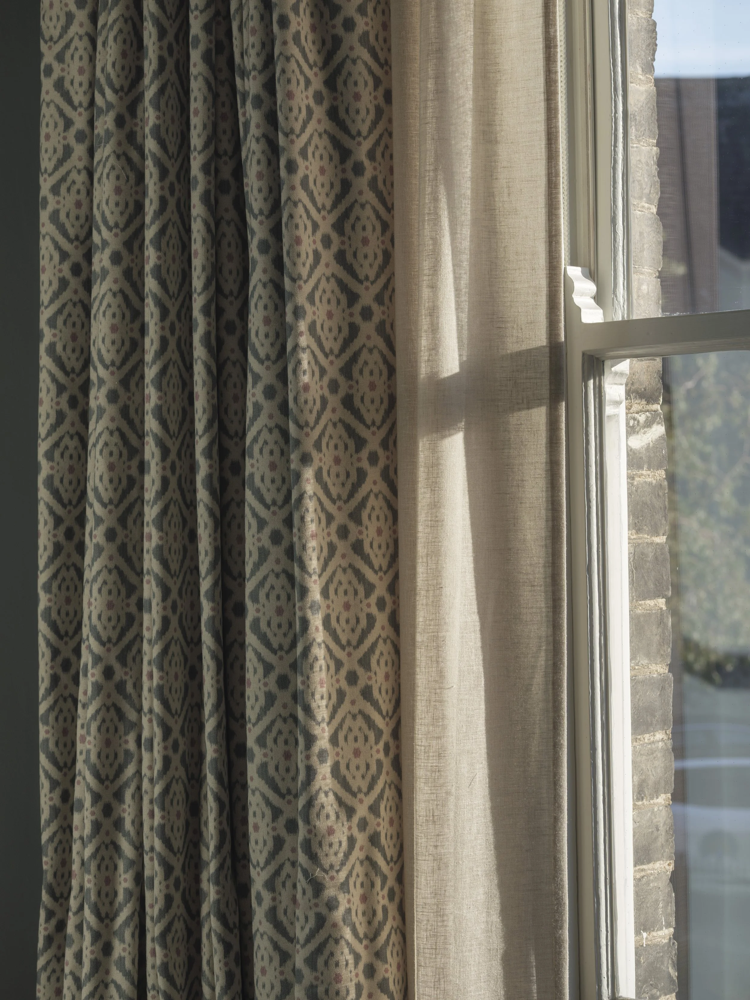 A window with beige sheer curtains and patterned drapes with geometric design, overlooking an exterior stone wall and a distant landscape.