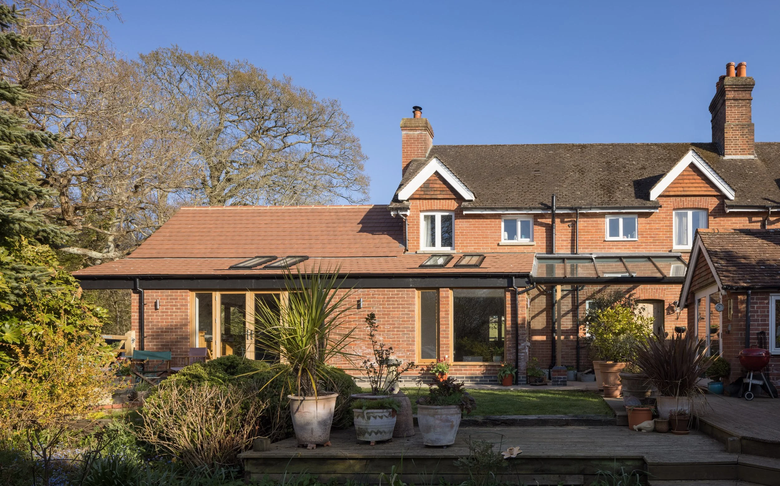 A residential house with a brick exterior, large windows, and a sloped roof. The backyard has a variety of potted plants, trees, and a wooden deck, under a clear blue sky.