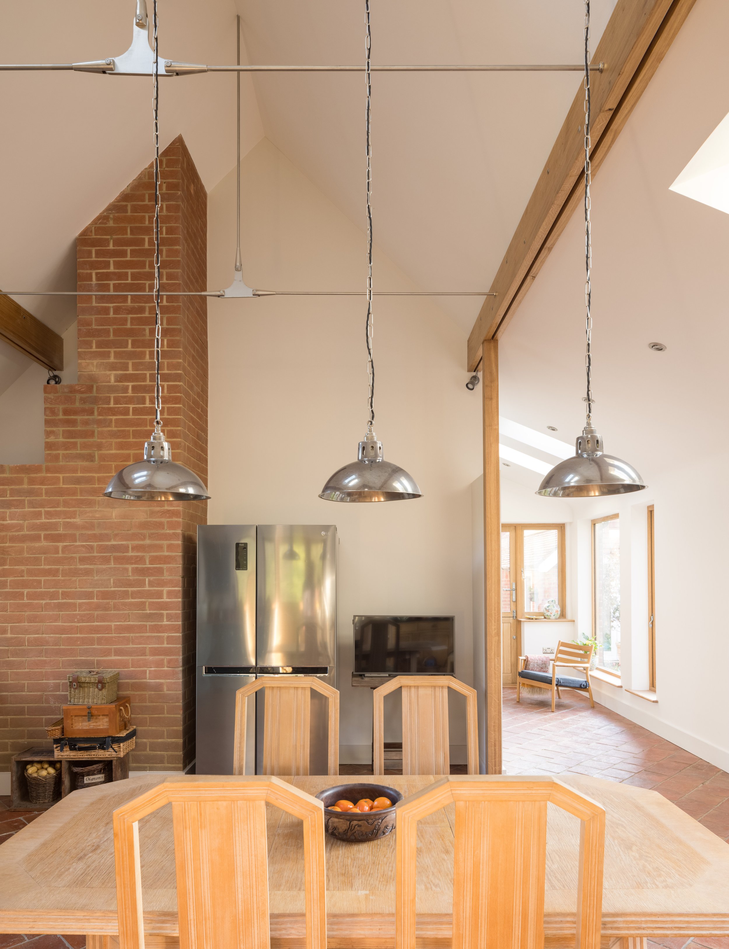 Interior view of a kitchen and dining area with a wooden dining table, four chairs, a bowl of tomatoes, a brick chimney, hanging industrial-style lights, and large windows allowing natural light.