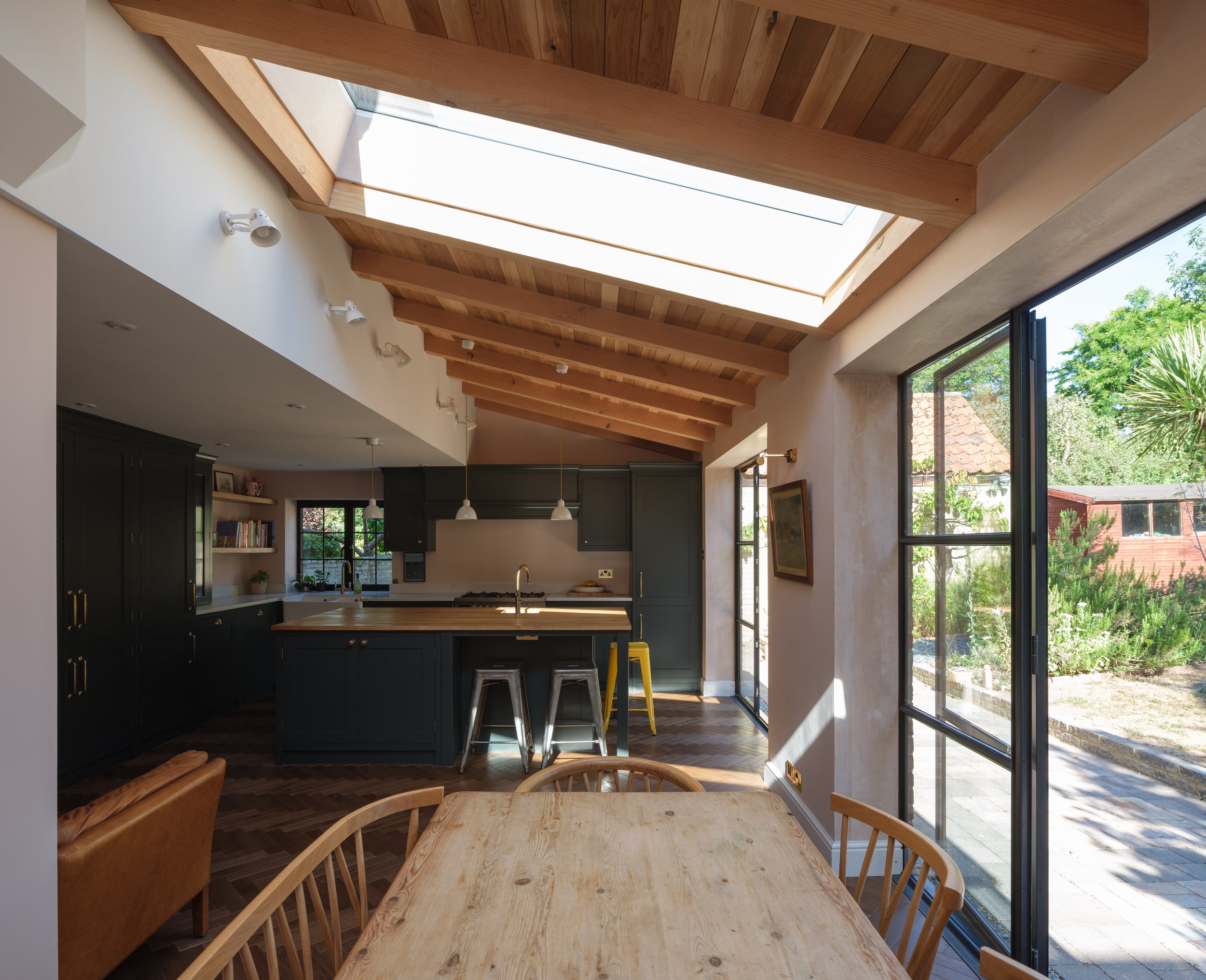 Interior view of a modern kitchen with a wooden ceiling, a large skylight, black cabinets, a kitchen island with bar stools, and a dining table with wooden chairs. Large windows reveal a garden outside.