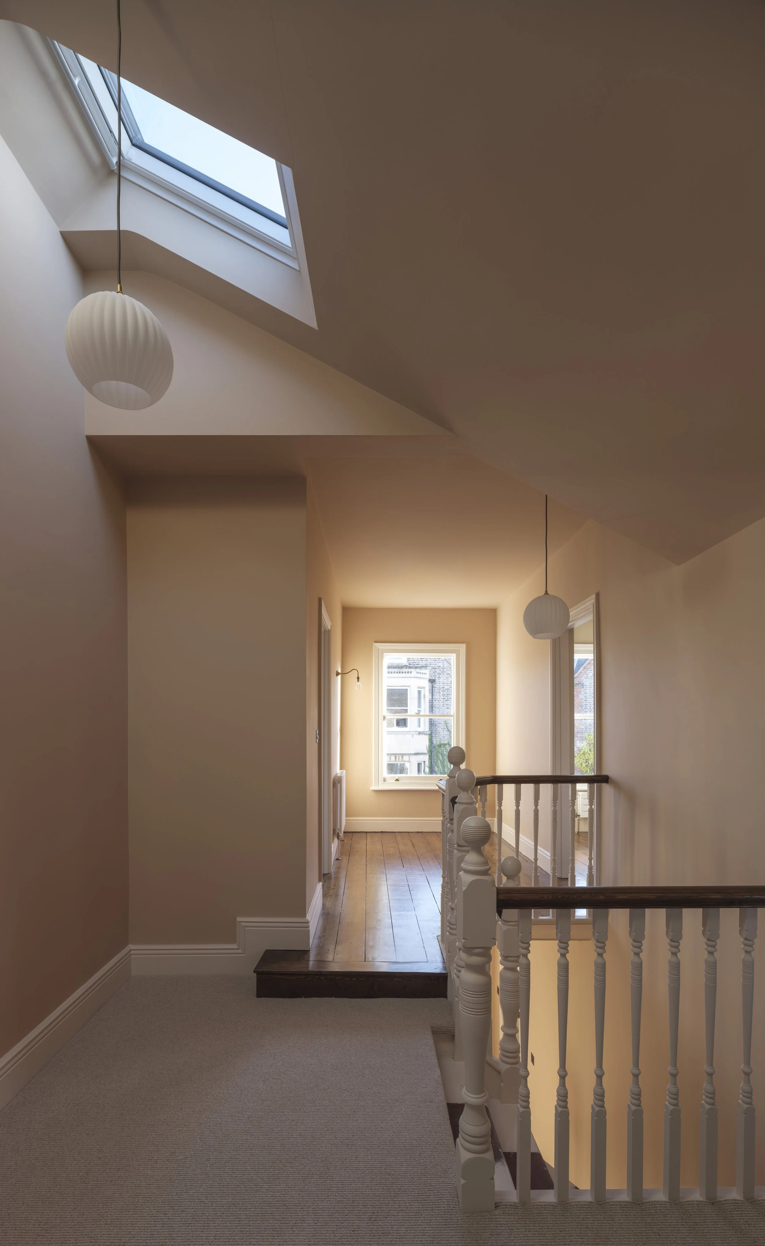 A hallway featuring a staircase with a white wooden railing, beige walls, and a beige carpeted floor. Natural light enters through a large window at the end of the hall, and a skylight above adds additional daylight. Two hanging pendant lights with w