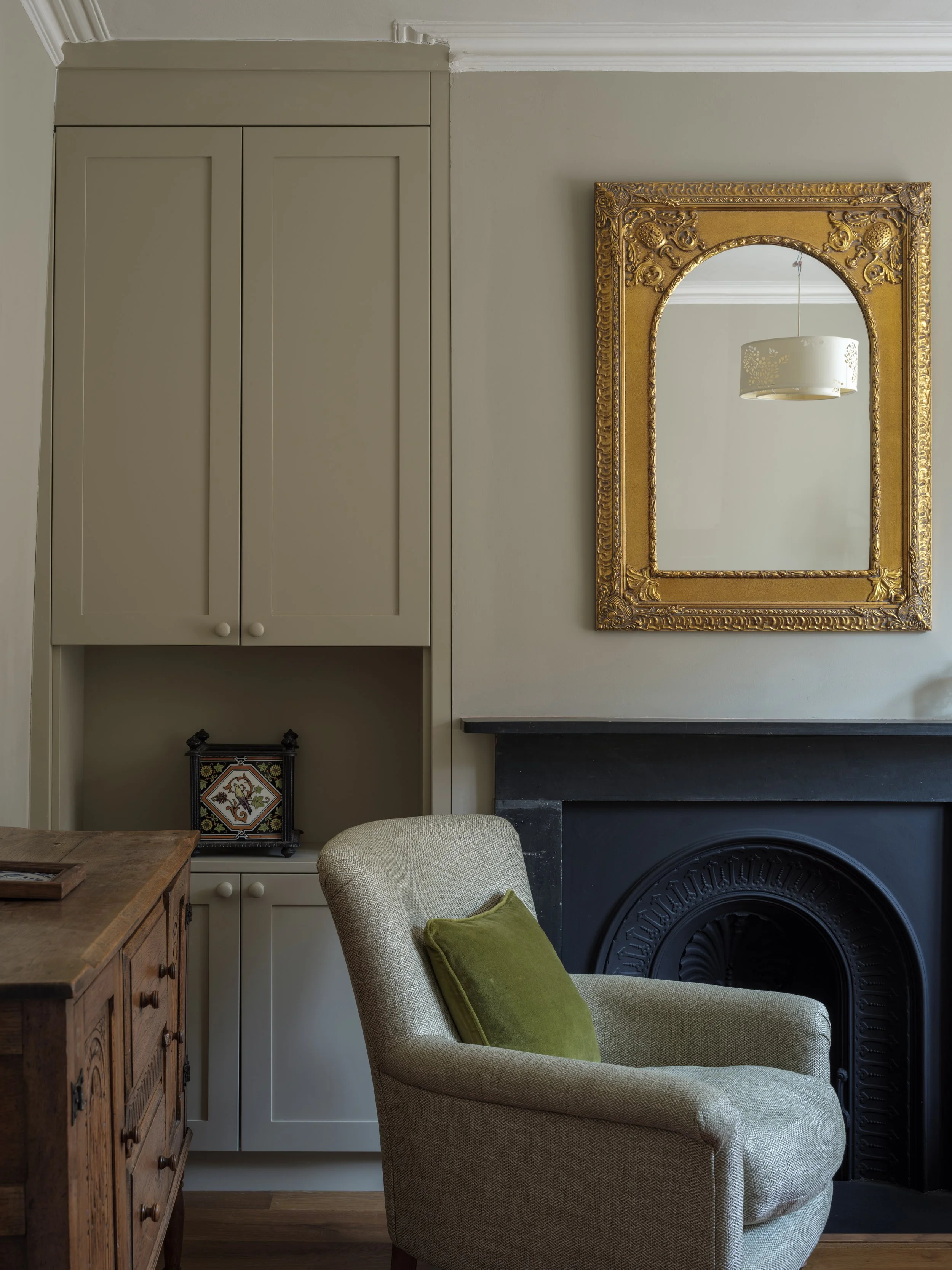 A cozy living room corner featuring an armchair with a green cushion, a wooden side table, a built-in cabinet, a black fireplace, a large ornate gold-framed mirror, and a paper lantern ceiling light reflected in the mirror.