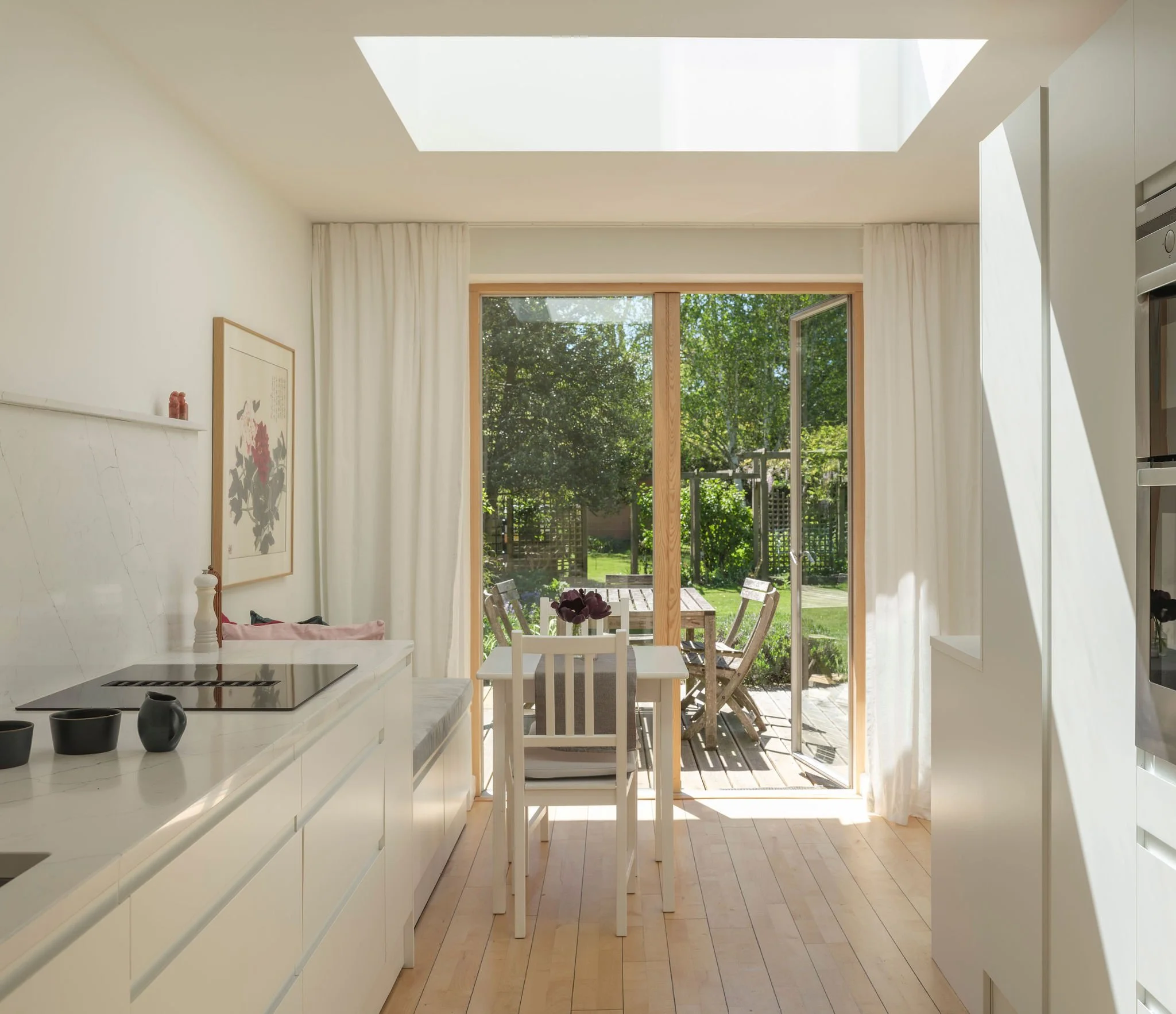 Sunlit kitchen area with a view through an open double door to an outdoor patio and lush green garden, featuring white cabinets, a marble countertop, and a dining table with chairs.