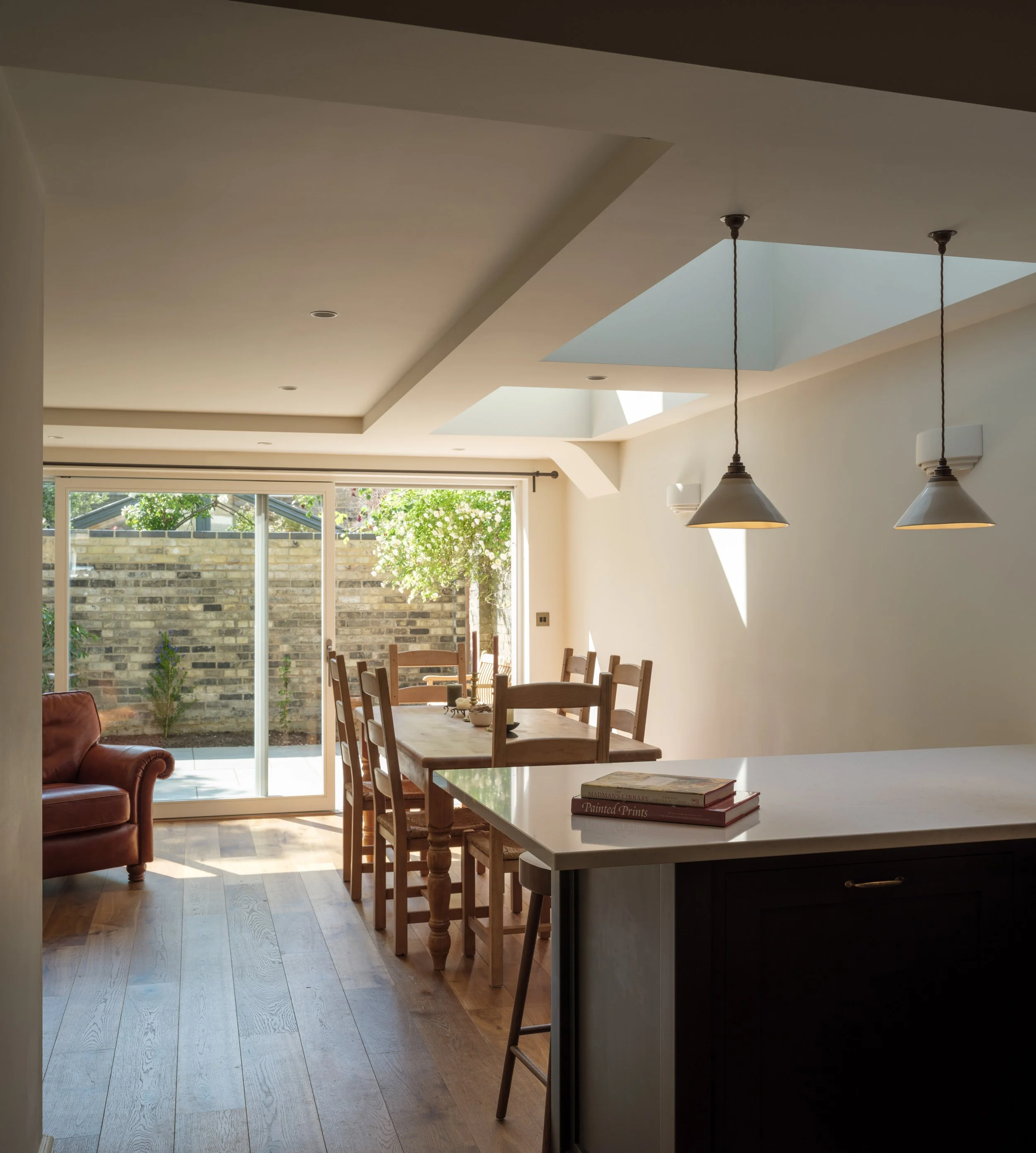 Bright dining room with wooden chairs around a table, a leather armchair in the corner, large sliding glass door opening to a backyard with a tree and brick wall, pendant lights hanging from the ceiling, and a clear skylight letting in natural light.