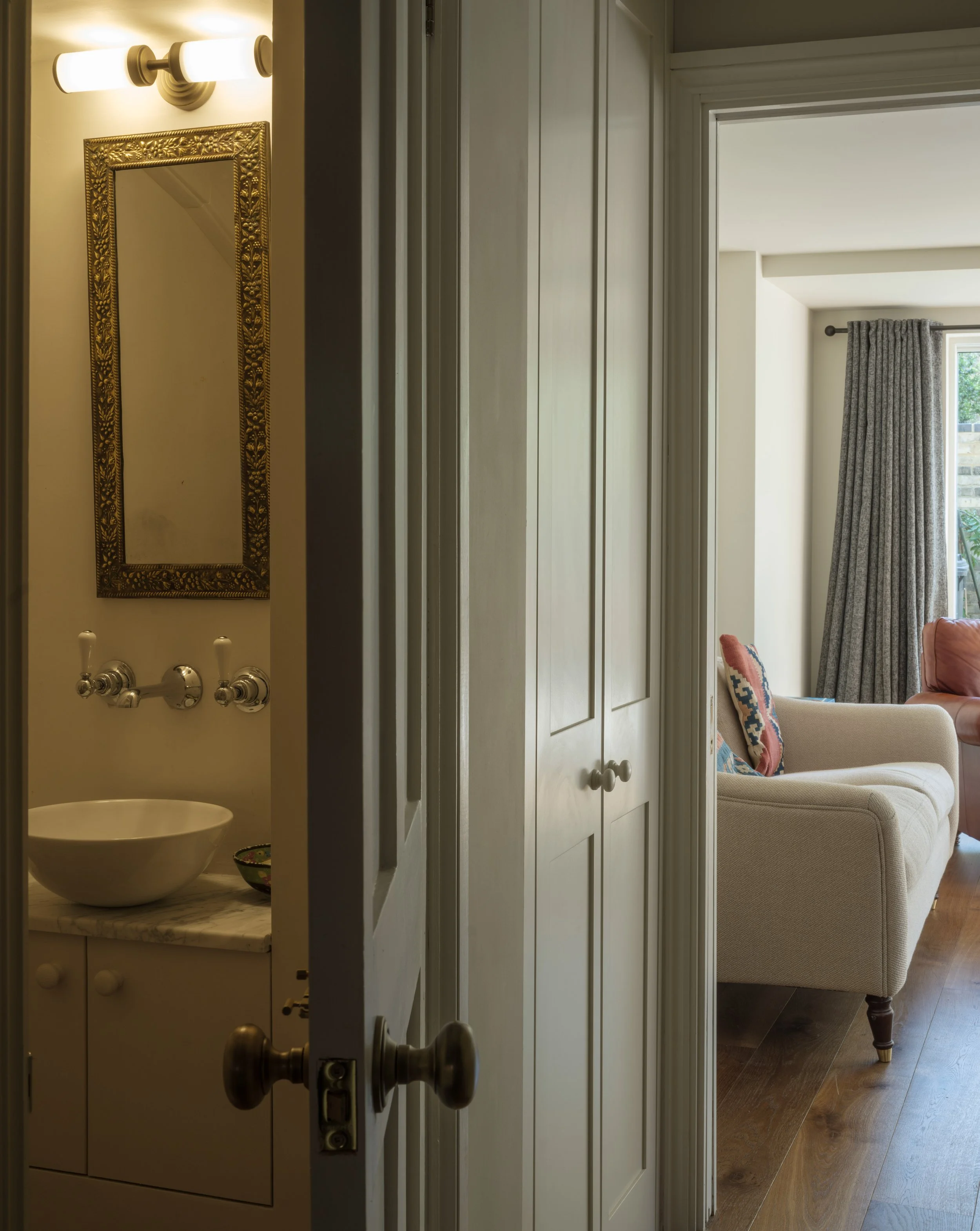 Partially open white door leading into a living room with beige walls, a window with gray curtains, a cream-colored sofa with patterned pillows, and hardwood floors.